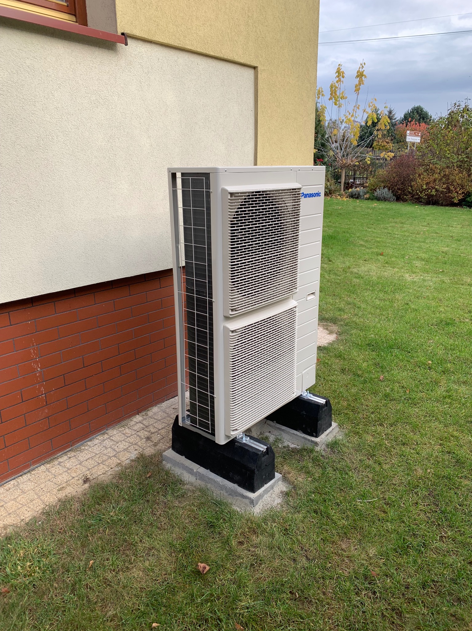Biała, dwuczęściowa pompa ciepła Panasonic zamontowana na czarnych podstawach przy ścianie budynku z ceglaną podmurówką i trawnikiem. Widok z boku, jesienna sceneria.