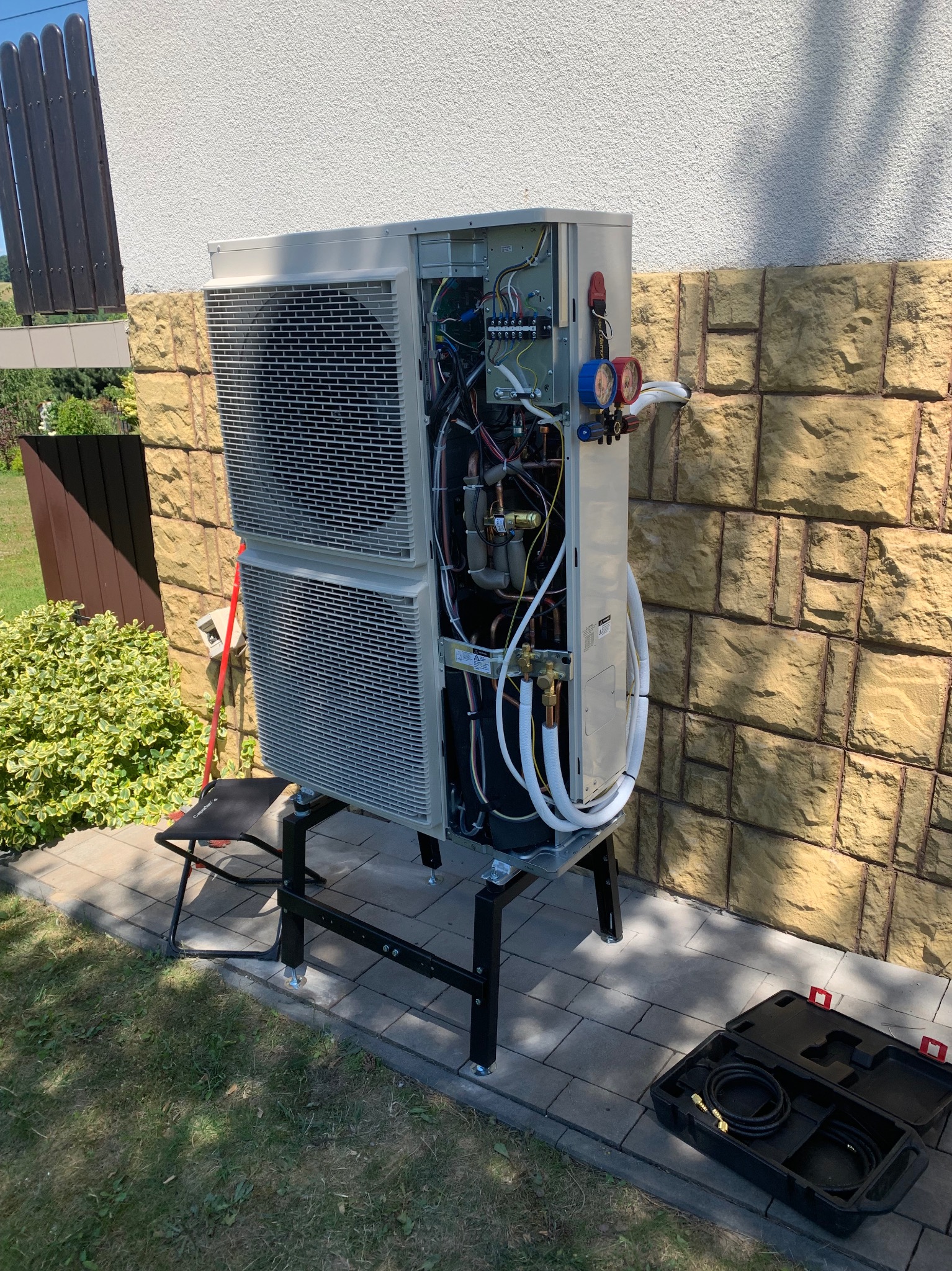 Otwarta obudowa dwuczęściowej pompy ciepła na zewnątrz budynku, widoczne instalacje, narzędzia i przyłącza. Obok mur z piaskowca i trawnik.