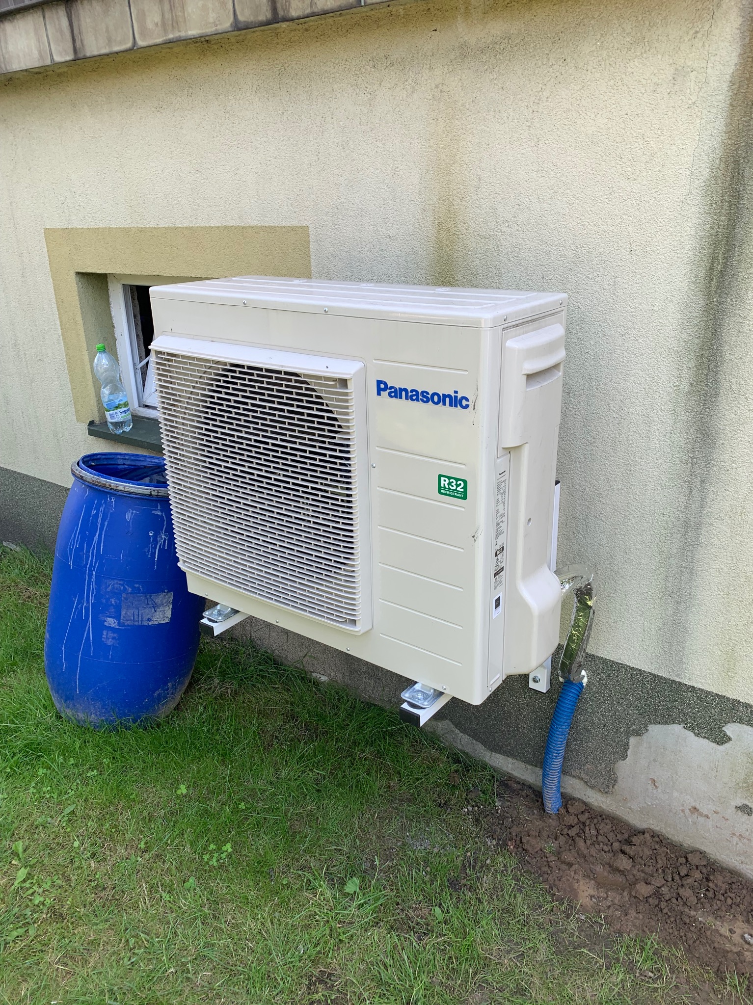 Zamontowany klimatyzator Panasonic na elewacji budynku w Częstochowie. Widoczna niebieska beczka obok urządzenia i fragment okna.
