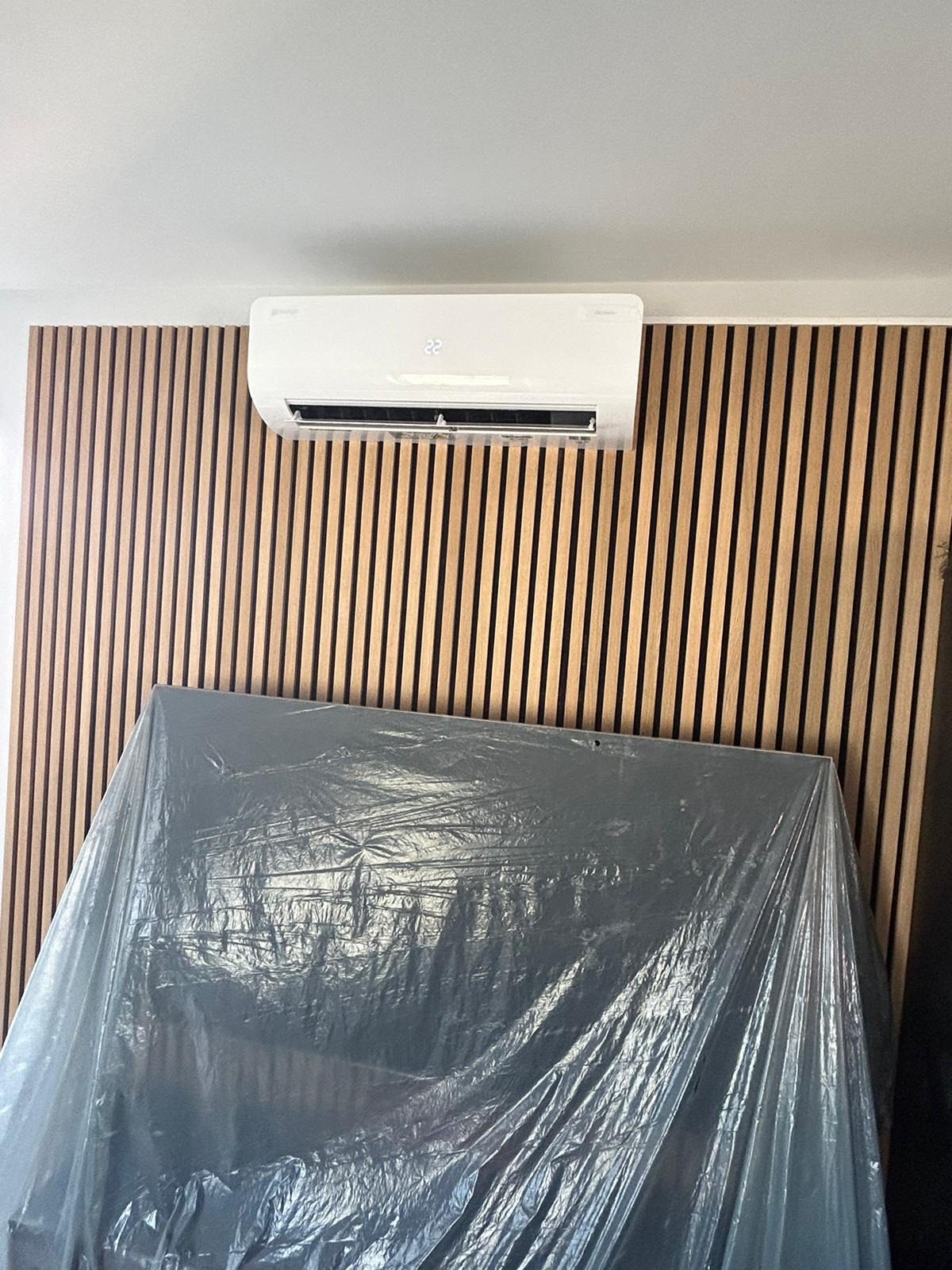Biała klimatyzacja na ścianie z drewnianych paneli, zabezpieczona folią meblową. Widoczny wyświetlacz temperatury na urządzeniu.