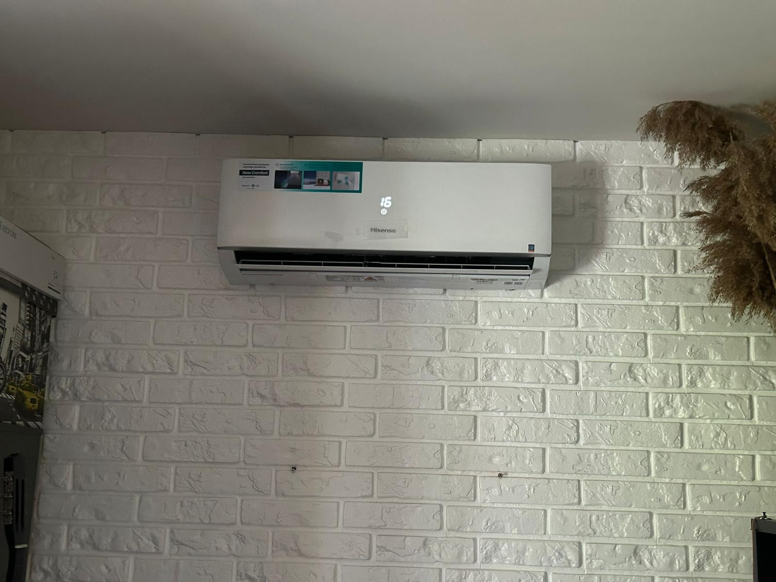 Biała klimatyzacja Hisense, zamontowana na ścianie z białej cegły. W tle widoczne dekoracyjne trawy pampasowe. Na wyświetlaczu urządzenia widoczna temperatura 16 stopni.