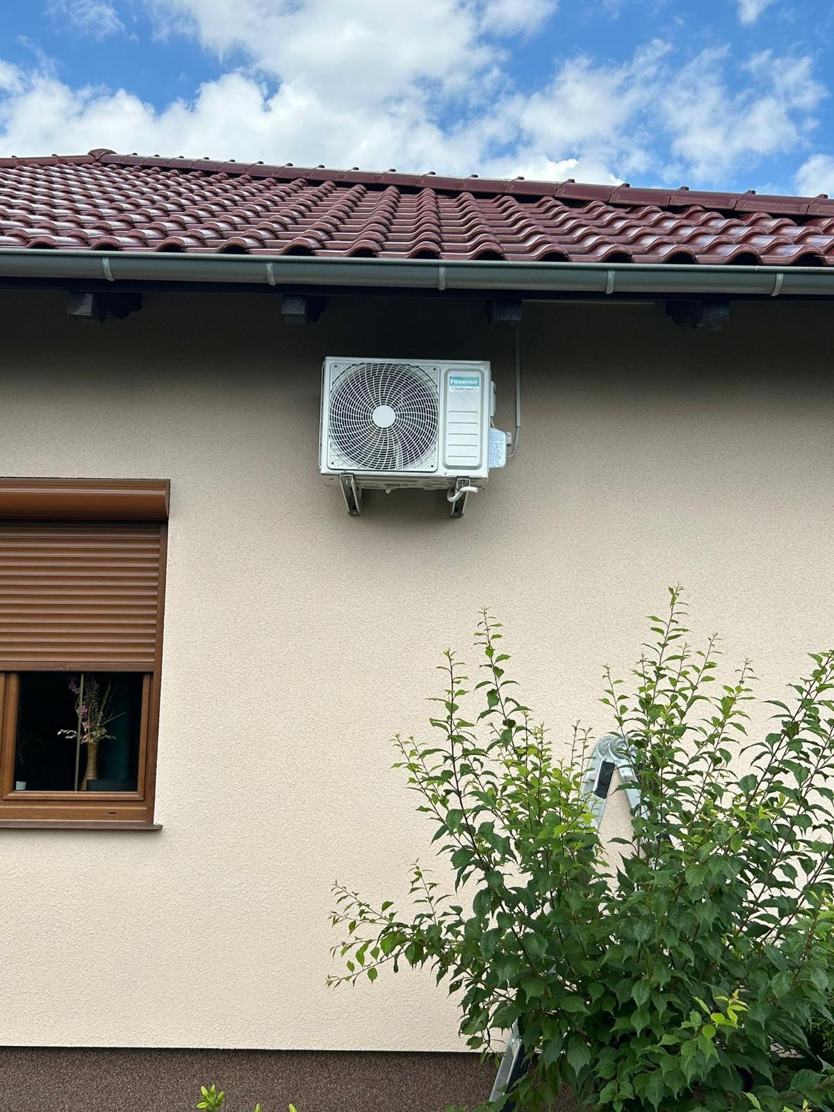 Biała klimatyzacja Hisense zamontowana na beżowej ścianie domu z brązowym dachem i oknem z roletą. Częściowo widoczna drabina i krzew. Błękitne niebo.