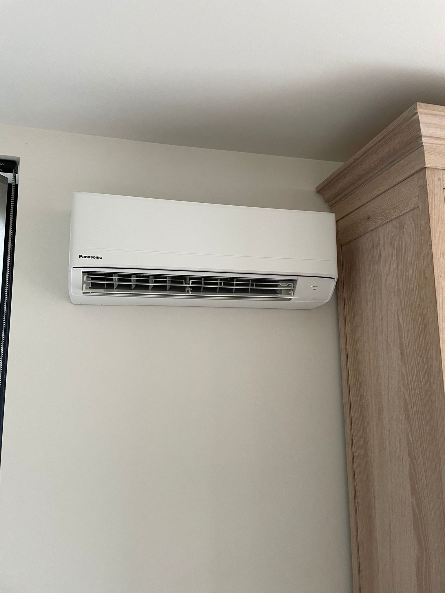 Biała klimatyzacja Panasonic na ścianie, z fragmentem drewnianej szafki po prawej. Neutralne tło, dobrze oświetlone wnętrze, minimalistyczny design.