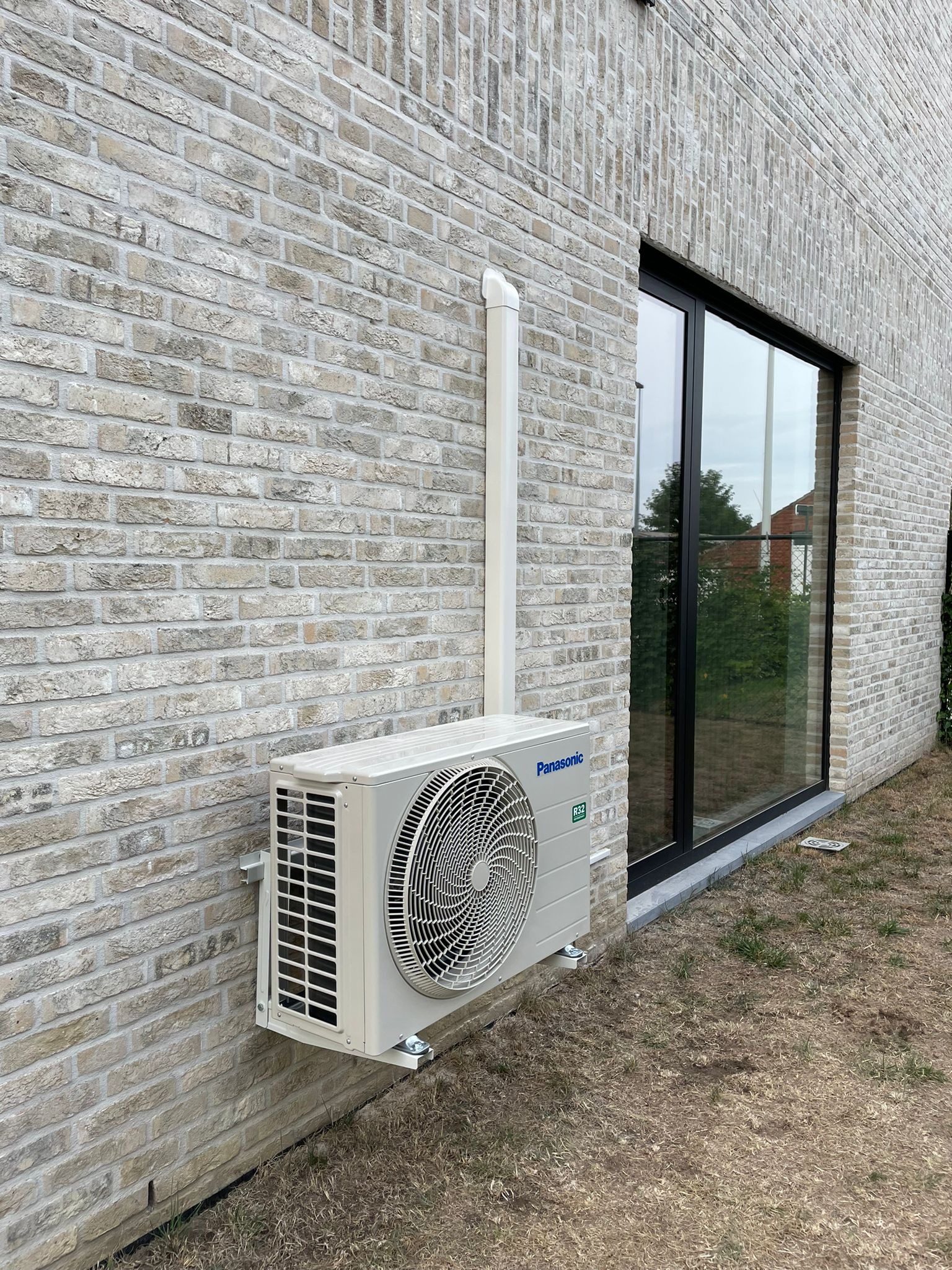 Zamontowana jednostka zewnętrzna klimatyzacji Panasonic na elewacji z cegły przy oknie. Widoczny biały przewód odprowadzający skropliny. Trawa przy ścianie.