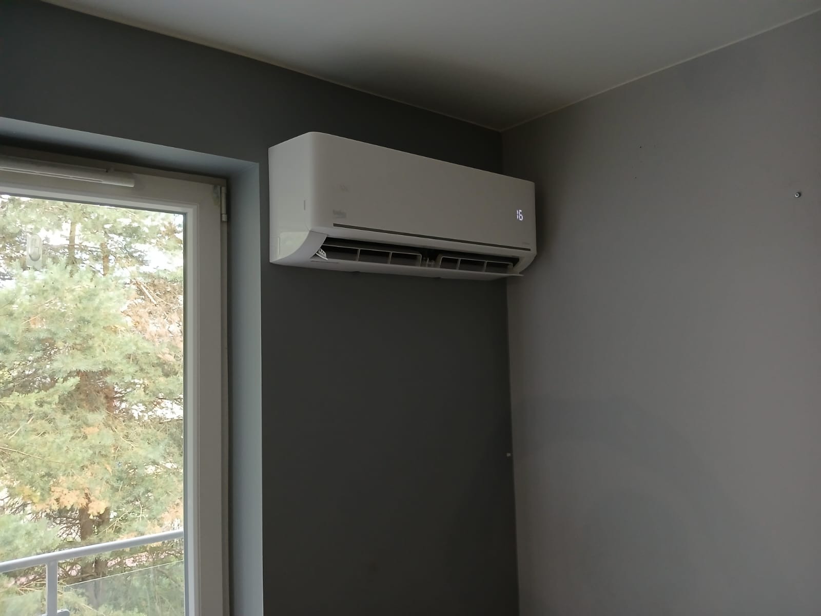 Biały klimatyzator na szarej ścianie w pokoju, widok z okna na zewnątrz. Na urządzeniu wyświetla się temperatura 16 stopni. Minimalistyczny design.