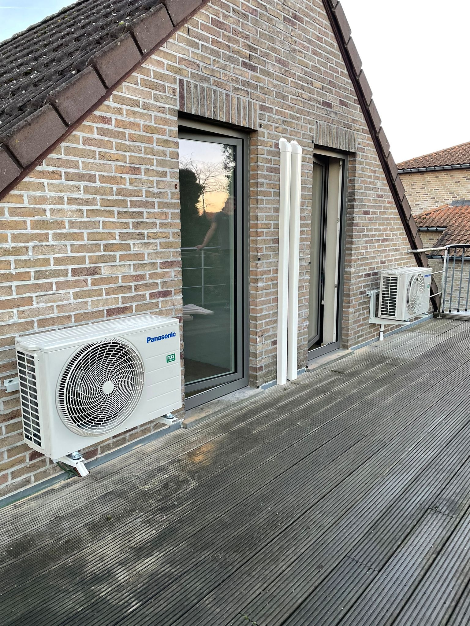 Zewnętrzna jednostka klimatyzacji Panasonic na balkonie z ceglaną ścianą i drewnianą podłogą. Widoczne rury odprowadzające. Ujęcie z niskiego kąta.