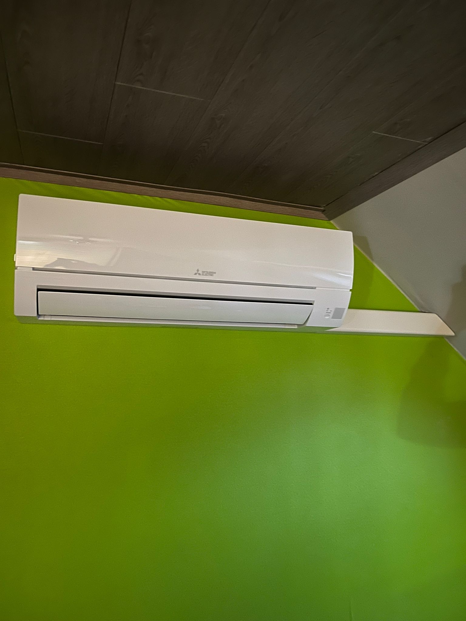 Biały klimatyzator Mitsubishi Electric zamontowany na zielonej ścianie w pomieszczeniu z ciemnym, drewnianym sufitem.