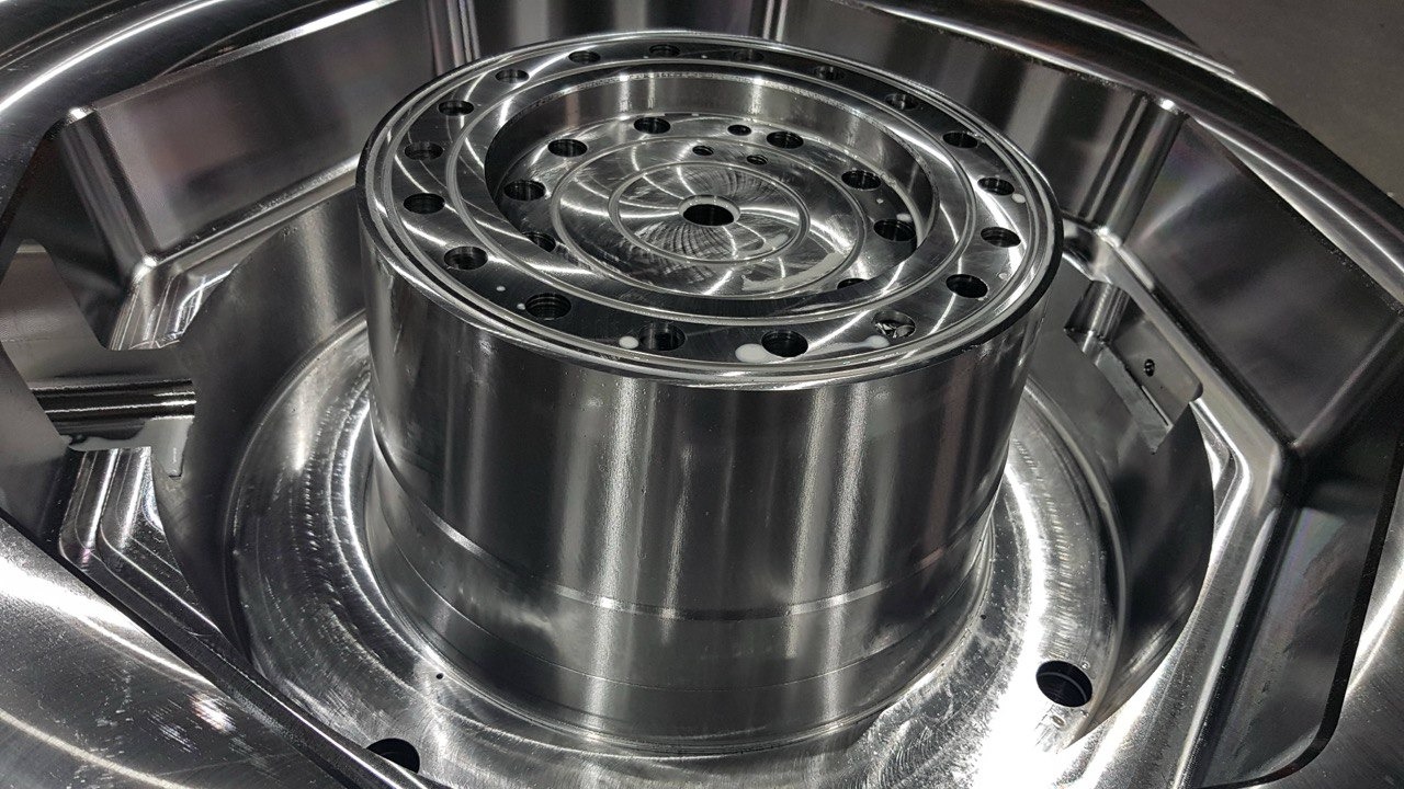 Precyzyjnie obrobiony, błyszczący, metalowy element z licznymi otworami i spiralnym wzorem, umieszczony wewnątrz większej, wyfrezowanej formy. Widoczne ślady chłodziwa.