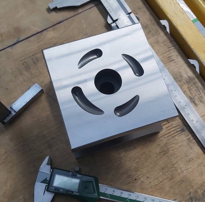 Precyzyjnie obrobiony, srebrny element sześcienny z okrągłym otworem centralnym i czterema wycięciami, mierzony suwmiarką na drewnianym stole.