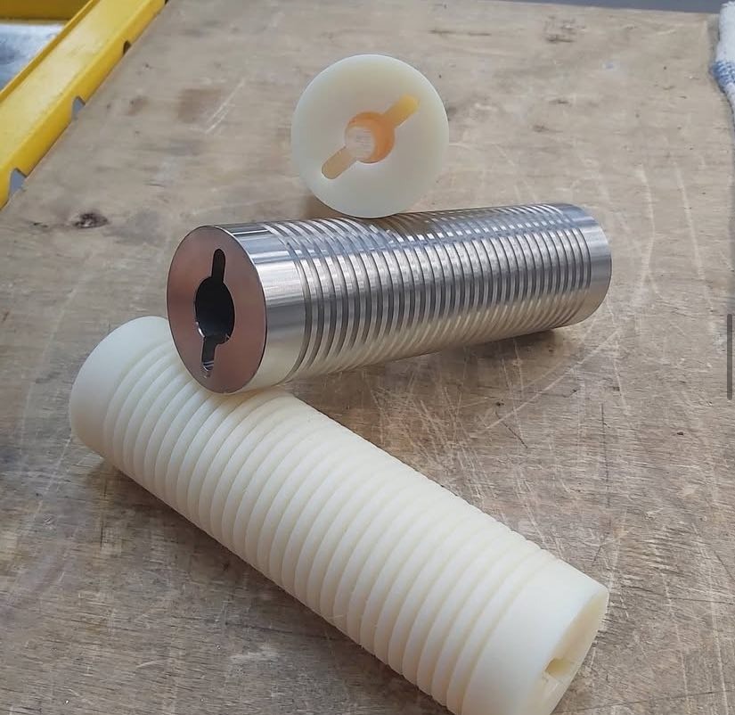 Precyzyjnie obrobione, cylindryczne elementy z metalu i tworzywa sztucznego z rowkami, leżące na drewnianym blacie. Widoczne detale obróbki.