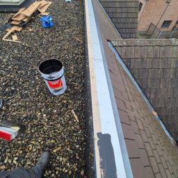 Gracjan Skalski - Prace na dachu pokrytym drobnymi kamieniami: malowanie blacharskich obróbek, widoczny kubeł z farbą, narzędzia, fragment dachu z dachówki.