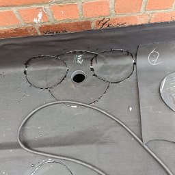 Gracjan Skalski - Przygotowanie membrany dachowej EPDM z wyciętymi otworami pod wpusty, widoczna ceglana ściana i narzędzia. Etap prac dekarskich, krycie dachu.