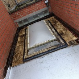 Gracjan Skalski - Płaski dach po renowacji w Buk. Widoczny świetlik dachowy i nowa, jasna hydroizolacja. Czerwona ściana budynku z cegły.