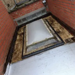 Płaski dach po renowacji w Buk. Widoczny świetlik dachowy i nowa, jasna hydroizolacja. Czerwona ściana budynku z cegły.