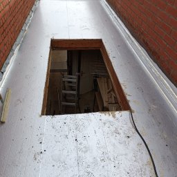 Gracjan Skalski - Płaski dach pokryty srebrną membraną z kwadratowym otworem, przez który widać drabinę i wnętrze. Ceglane ściany po bokach dachu.