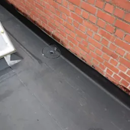 Płaski dach pokryty czarną membraną bitumiczną, przylegający do ceglanej ściany. Widoczny odpływ wody i pęknięcie na powierzchni dachu.