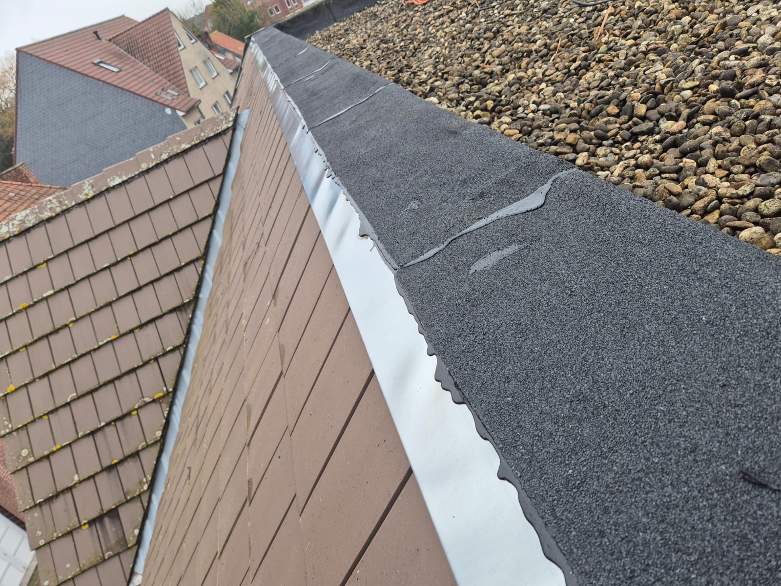 Szczegółowe ujęcie dachu z papy z posypką, obróbki blacharskiej i fragmentu dachu pokrytego dachówką. W tle widać inne budynki.