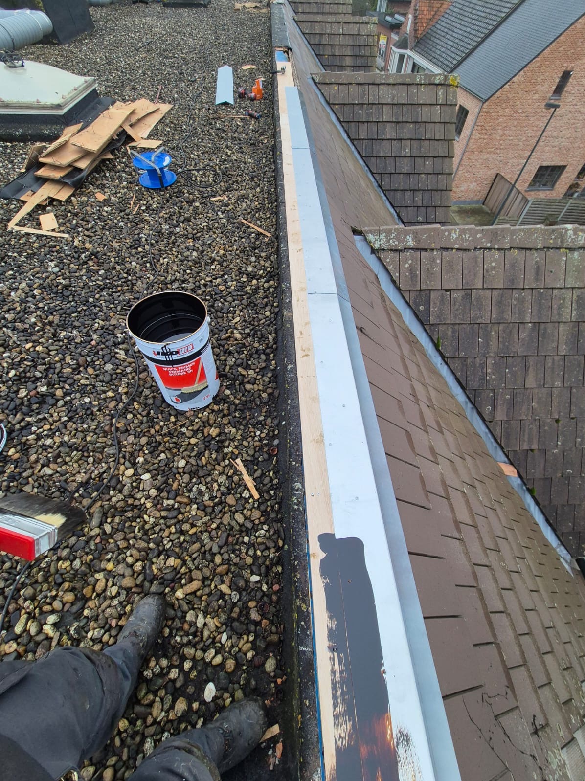Prace na dachu pokrytym drobnymi kamieniami: malowanie blacharskich obróbek, widoczny kubeł z farbą, narzędzia, fragment dachu z dachówki.