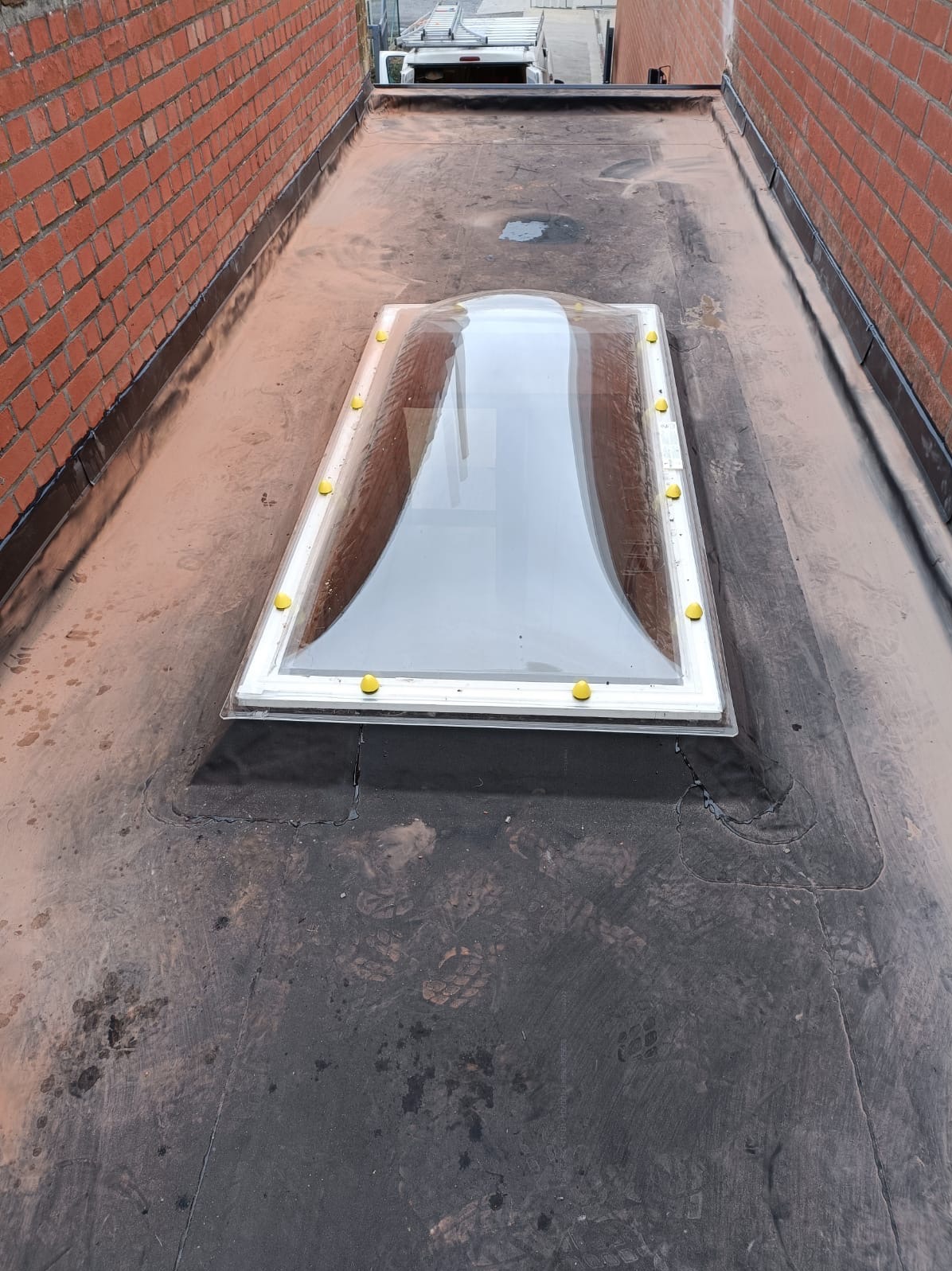 Płaski dach pokryty papą z zamontowanym świetlikiem. Widoczne ślady stóp na papie i ceglane ściany boczne. Na dachu żółte elementy mocujące świetlik.