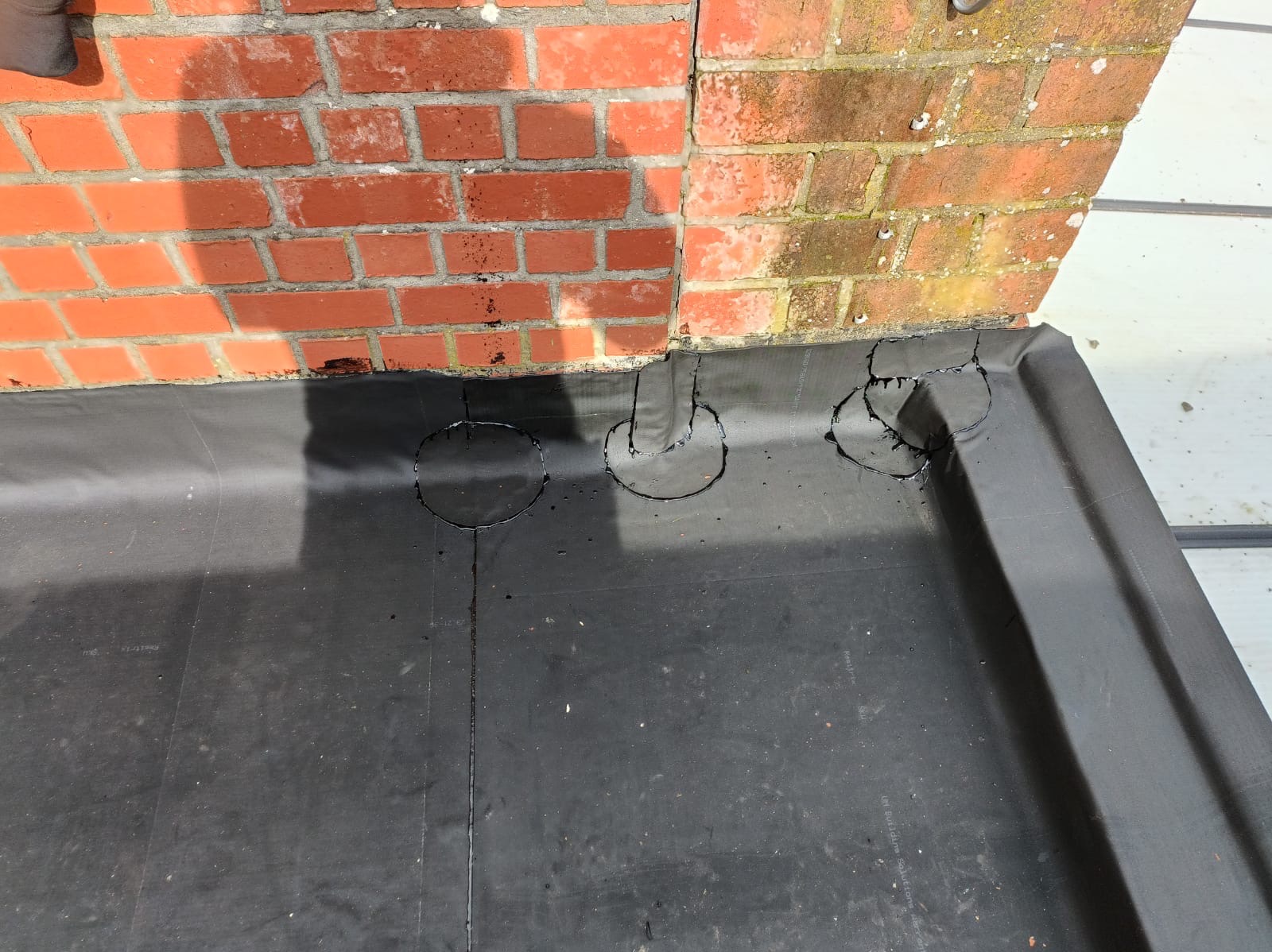 Uszczelnienie dachu papą bitumiczną wokół komina z cegły. Widoczne okrągłe ślady po obróbce detali. Szare deski w tle, minimalne nachylenie.