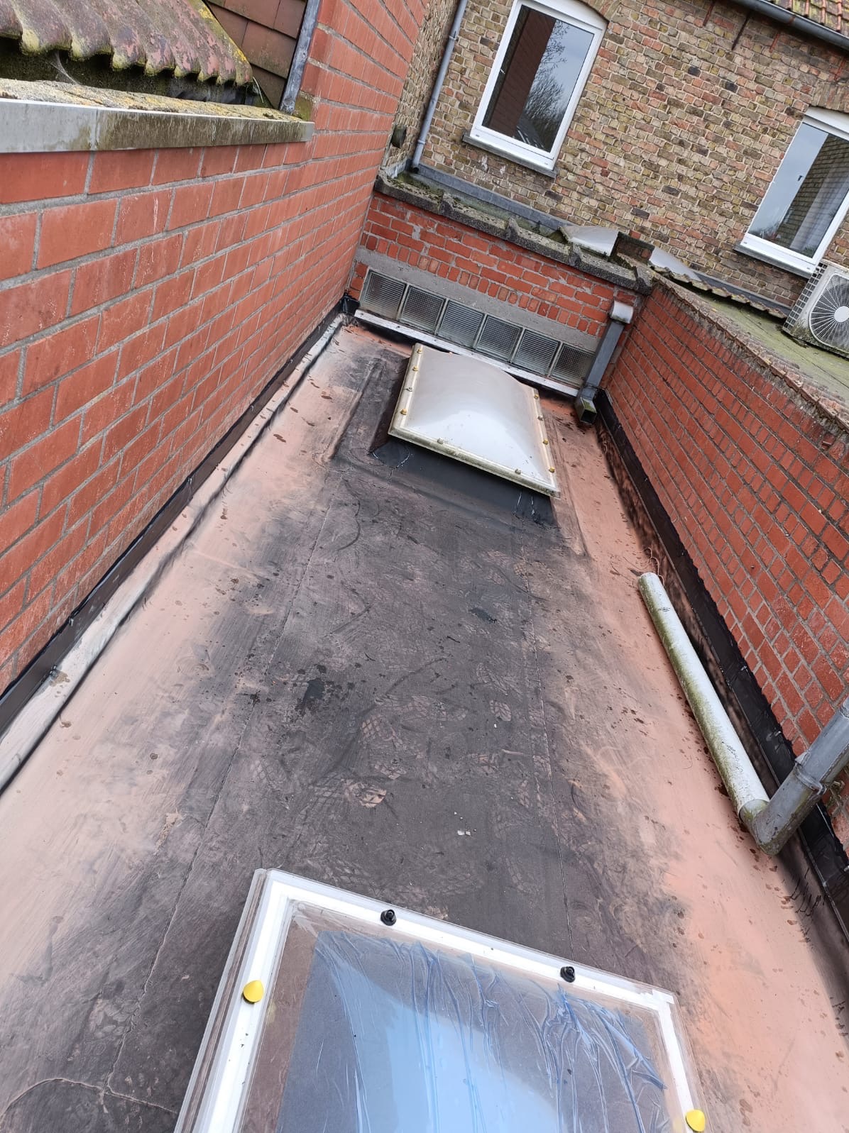 Płaski dach z bitumiczną membraną i dwoma świetlikami. Widoczne ślady użytkowania, ceglana ściana i rynna. Górny świetlik z uszkodzeniami.