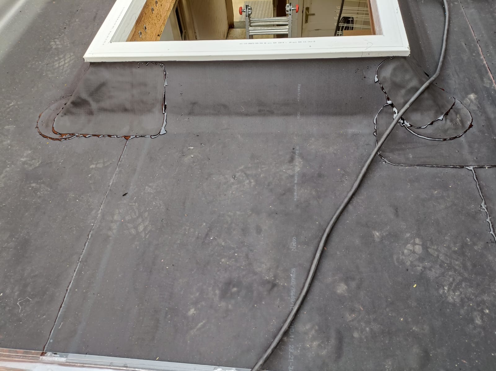 Uszczelnianie dachu z papy bitumicznej wokół okna dachowego, widoczne ślady po zalaniu i czarny kabel. Prace dekarskie w toku.