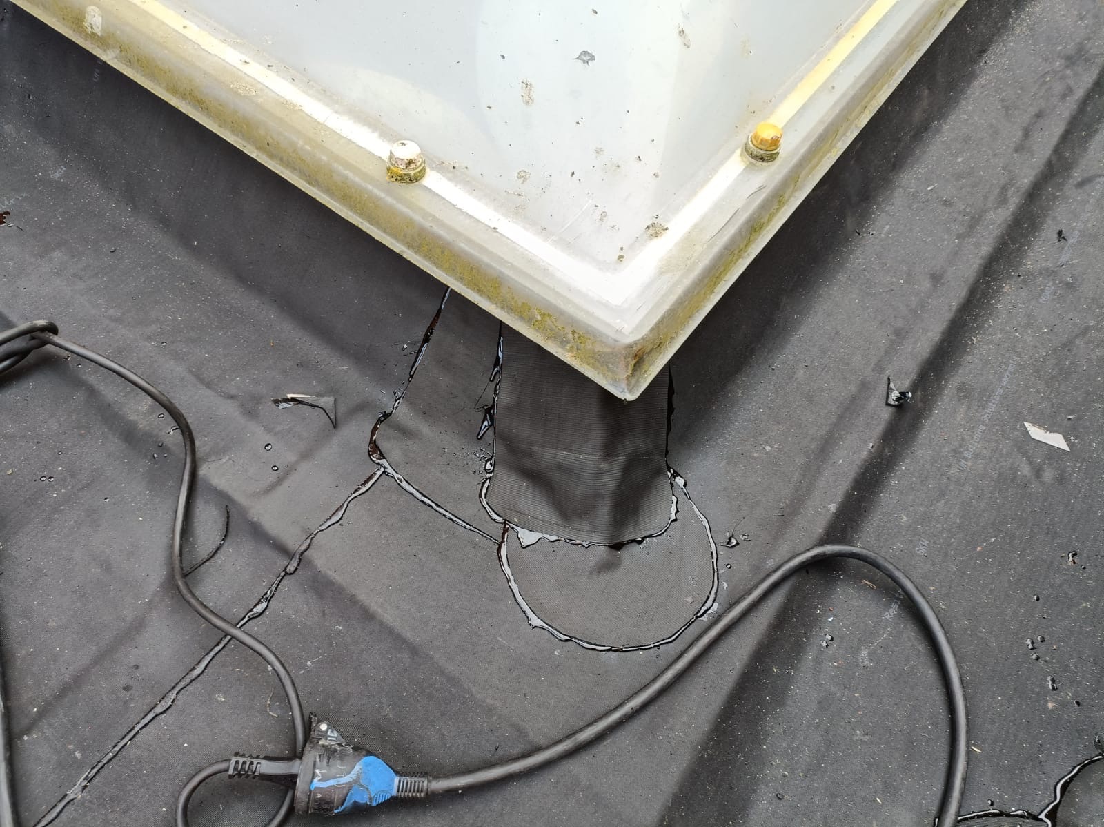 Fragment dachu z przeciekającym świetlikiem i kablem elektrycznym. Widoczne ślady wilgoci na czarnej membranie dachowej.