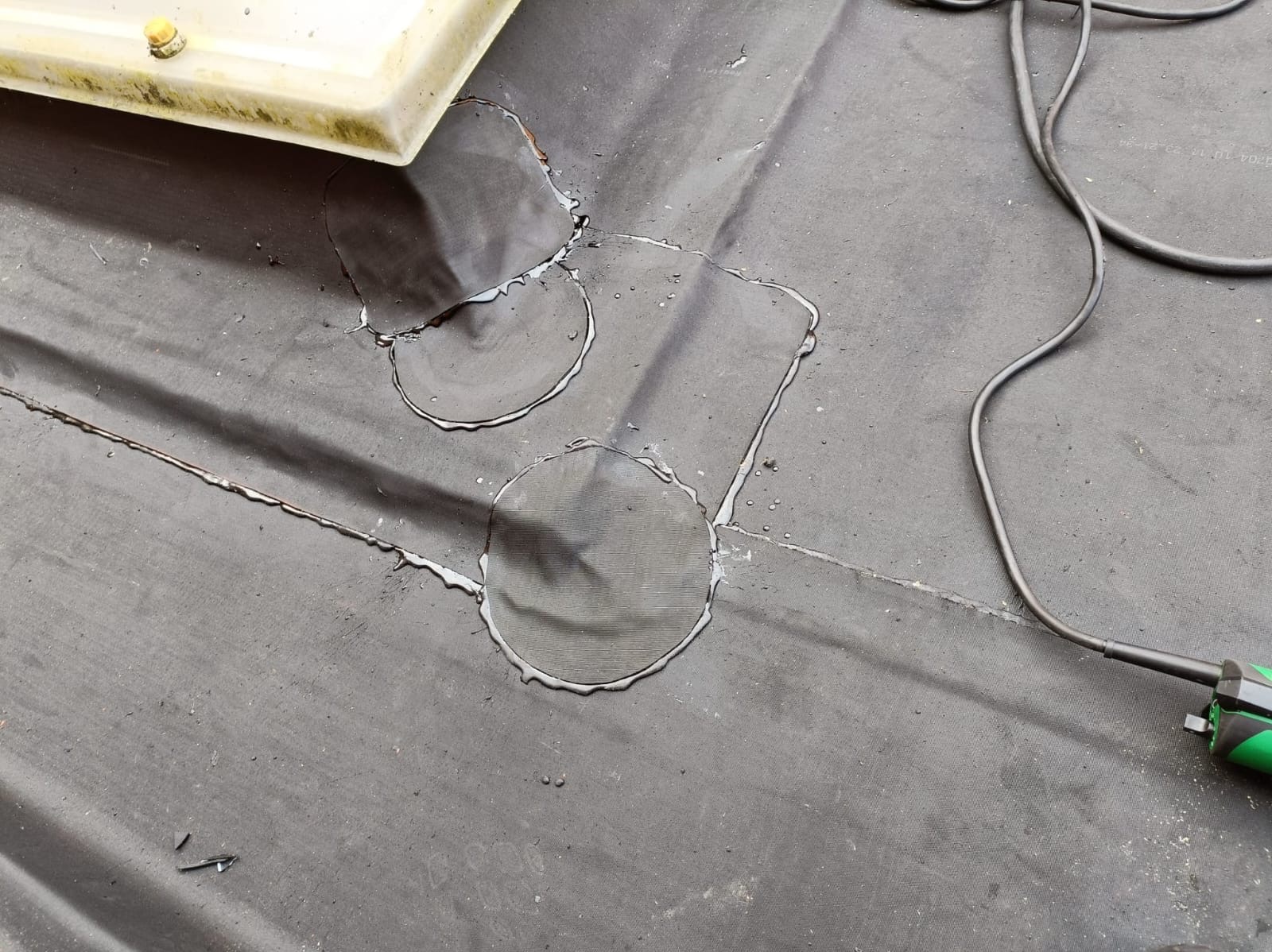 Szczegółowe ujęcie załatanych miejsc na czarnym pokryciu dachowym, z widocznym narzędziem i elementem instalacji. Widoczne ślady kleju i napraw.