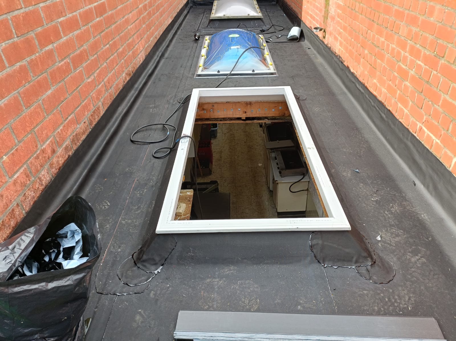 Czarny dach pokryty papą z otworem okiennym i świetlikami, widok z góry. Na dachu leżą narzędzia i odpady. Mur z czerwonej cegły w tle.