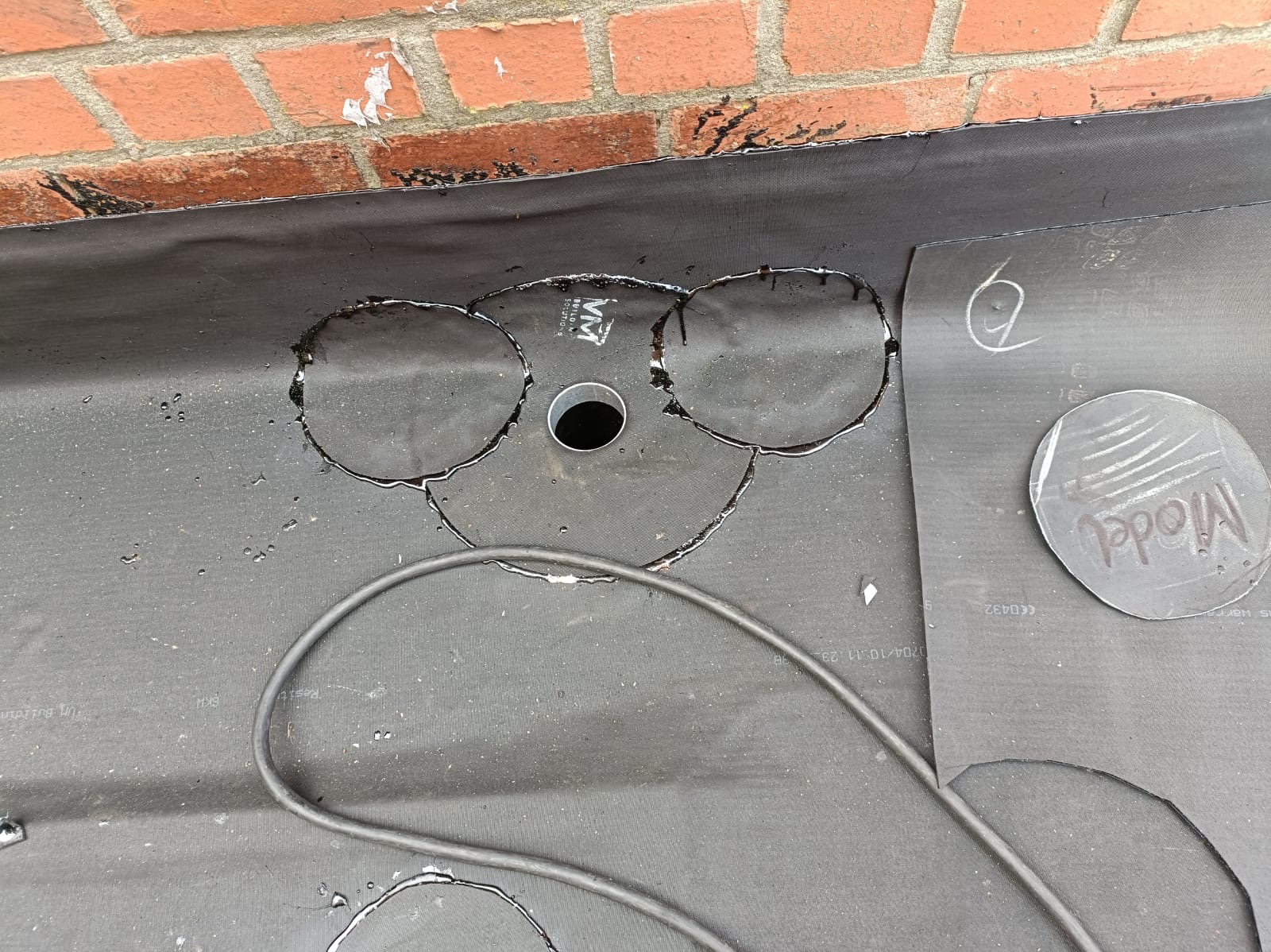 Przygotowanie membrany dachowej EPDM z wyciętymi otworami pod wpusty, widoczna ceglana ściana i narzędzia. Etap prac dekarskich, krycie dachu.