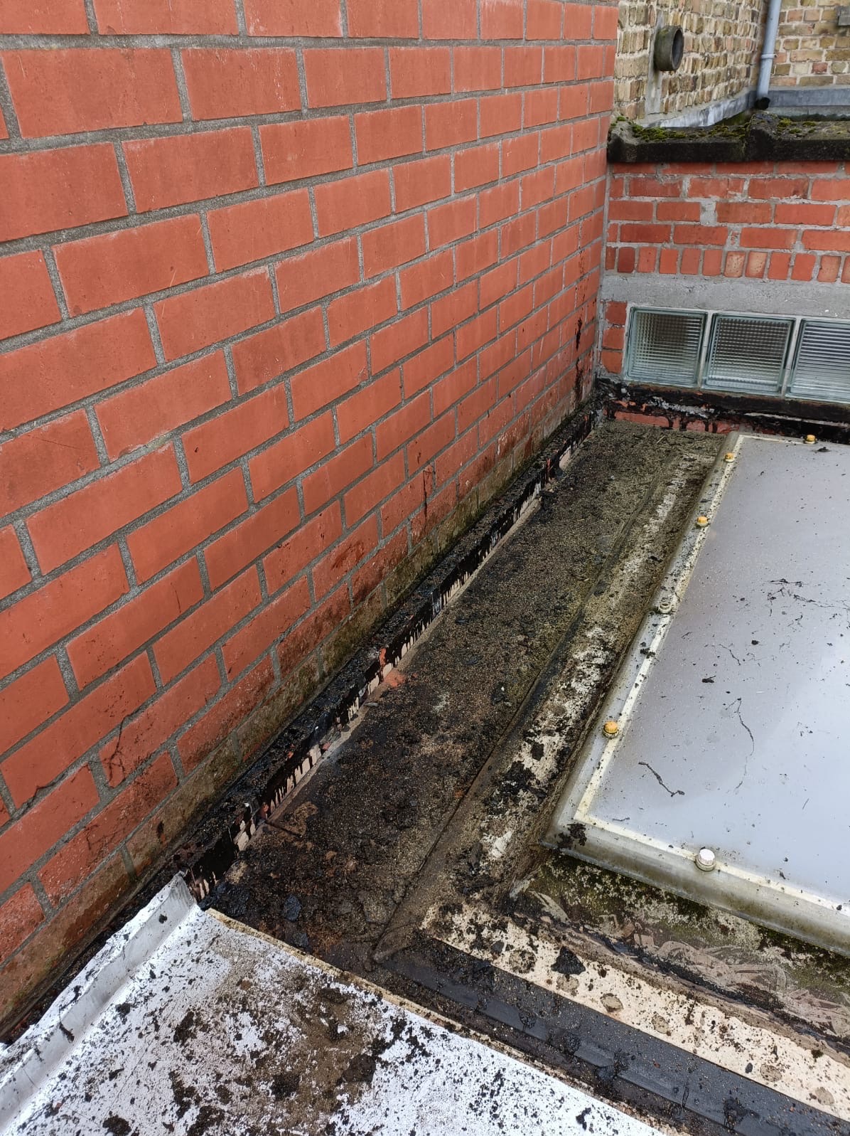 Nieszczelny, płaski dach z bitumiczną powłoką przy ceglanej ścianie. Widoczne ślady przecieków i zabrudzenia na dachu i ścianie, co sugeruje potrzebę naprawy.