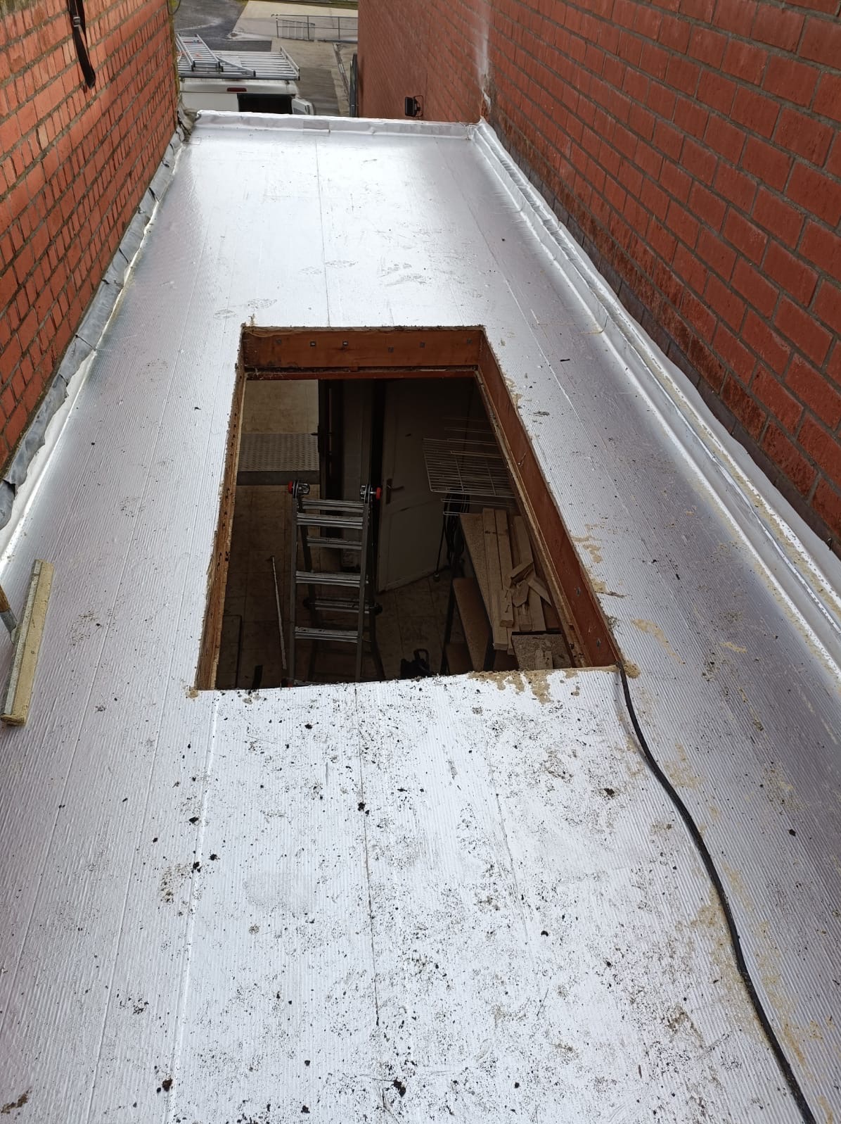 Płaski dach pokryty srebrną membraną z kwadratowym otworem, przez który widać drabinę i wnętrze. Ceglane ściany po bokach dachu.