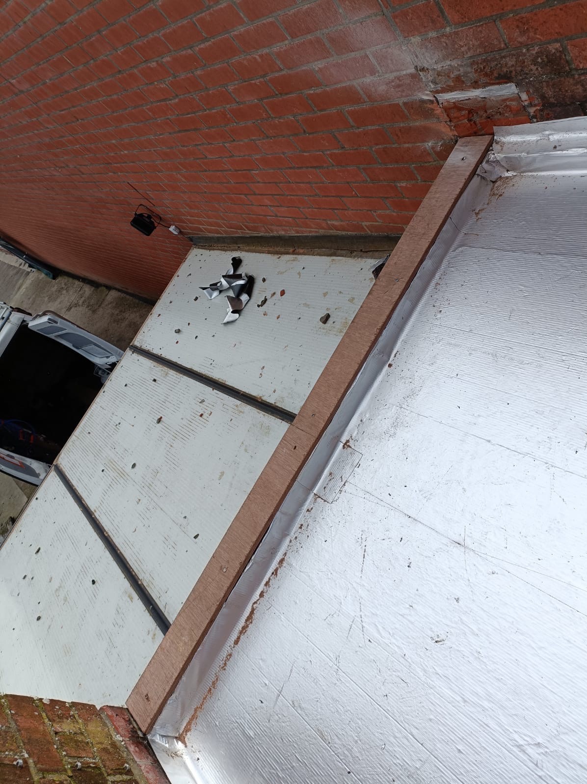 Fragment dachu w trakcie prac: drewniana listwa przy krawędzi, izolacja termiczna, widoczny fragment ściany z cegły i narzędzia. Ujęcie z góry, pod kątem.