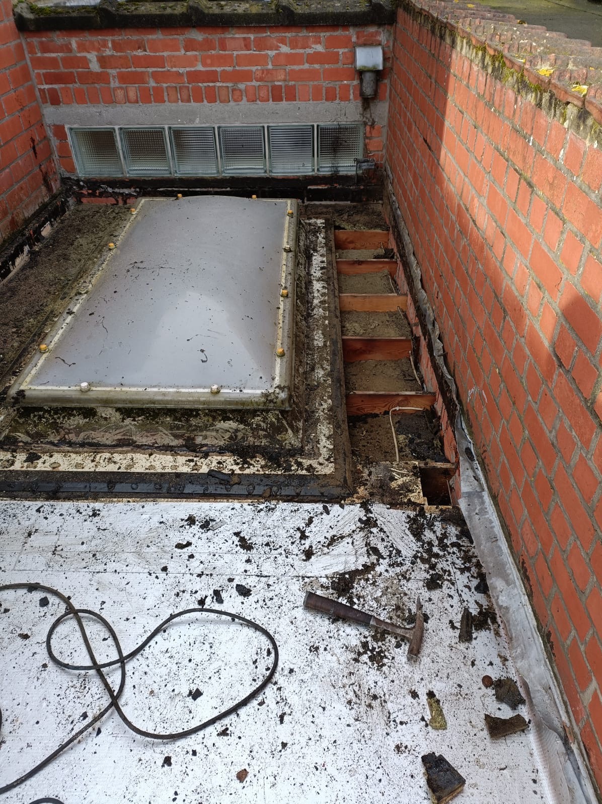 Odkryty fragment dachu z oknem dachowym i odsłoniętą konstrukcją drewnianą, ceglana ściana, widoczne narzędzia i resztki materiałów budowlanych.