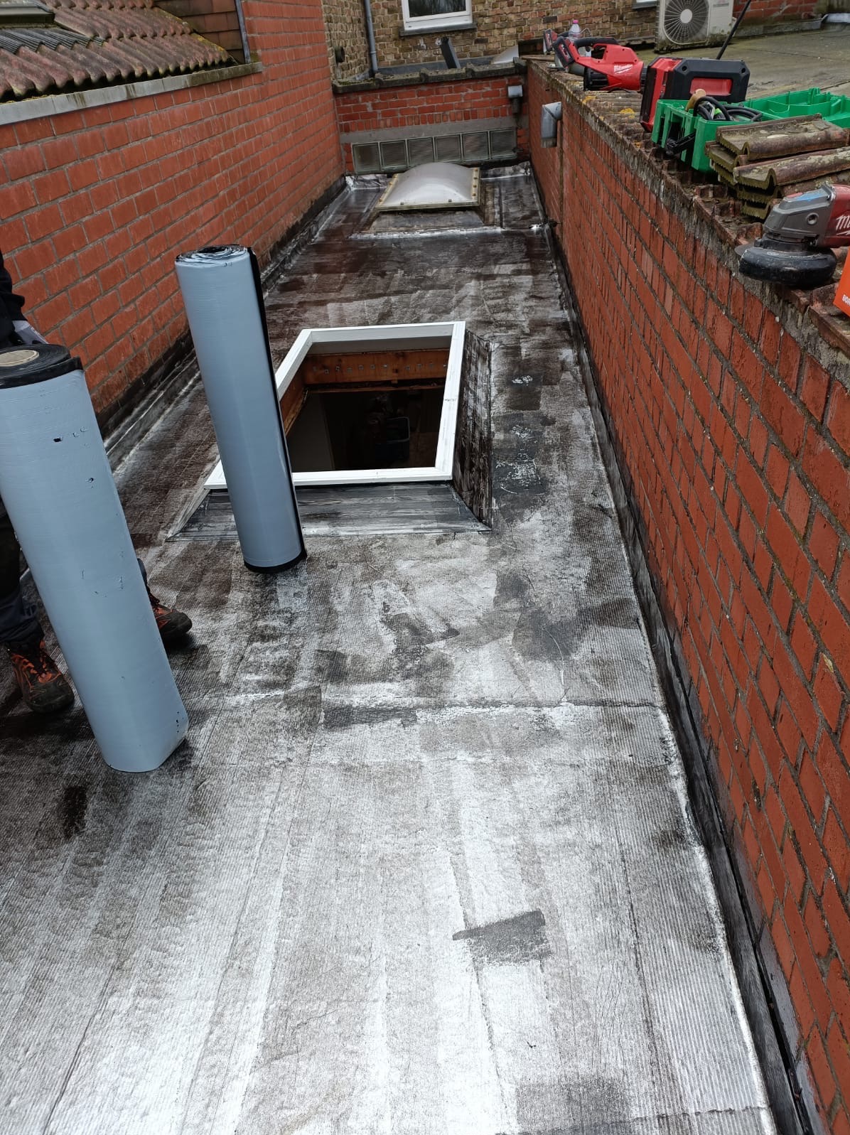 Prace dekarskie na płaskim dachu: papa bitumiczna, świetlik dachowy, narzędzia. Widok z góry na częściowo pokryty dach z dwoma rolkami papy.