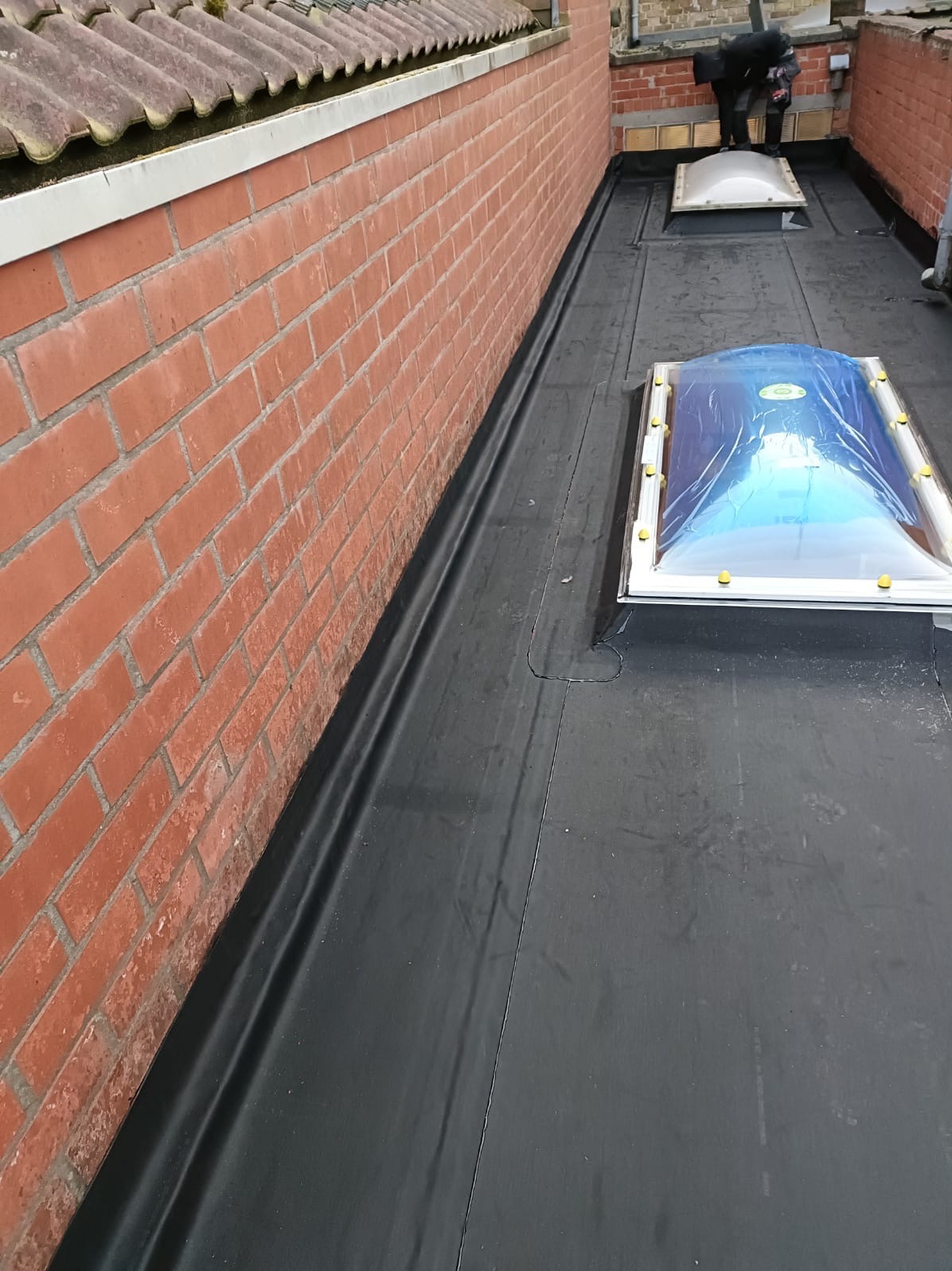 Wykończony, płaski dach pokryty czarną membraną, z dwoma świetlikami (jeden z folią ochronną). Czerwona ceglana ściana z boku. Prace dekarskie w tle, widoczna osoba.