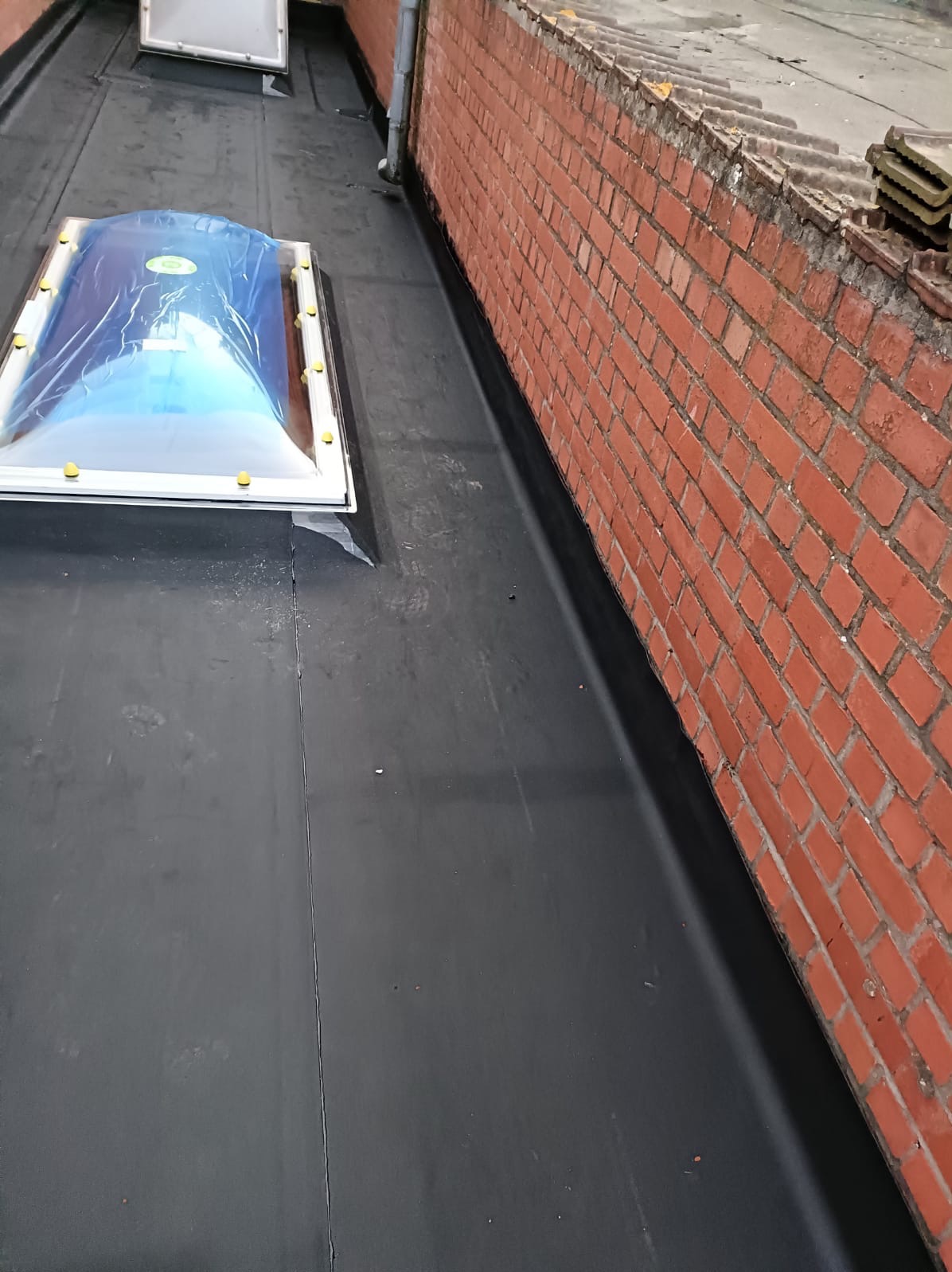 Płaski dach pokryty czarną papą, z zamontowanym świetlikiem zabezpieczonym folią. Mur z czerwonej cegły wzdłuż krawędzi dachu.