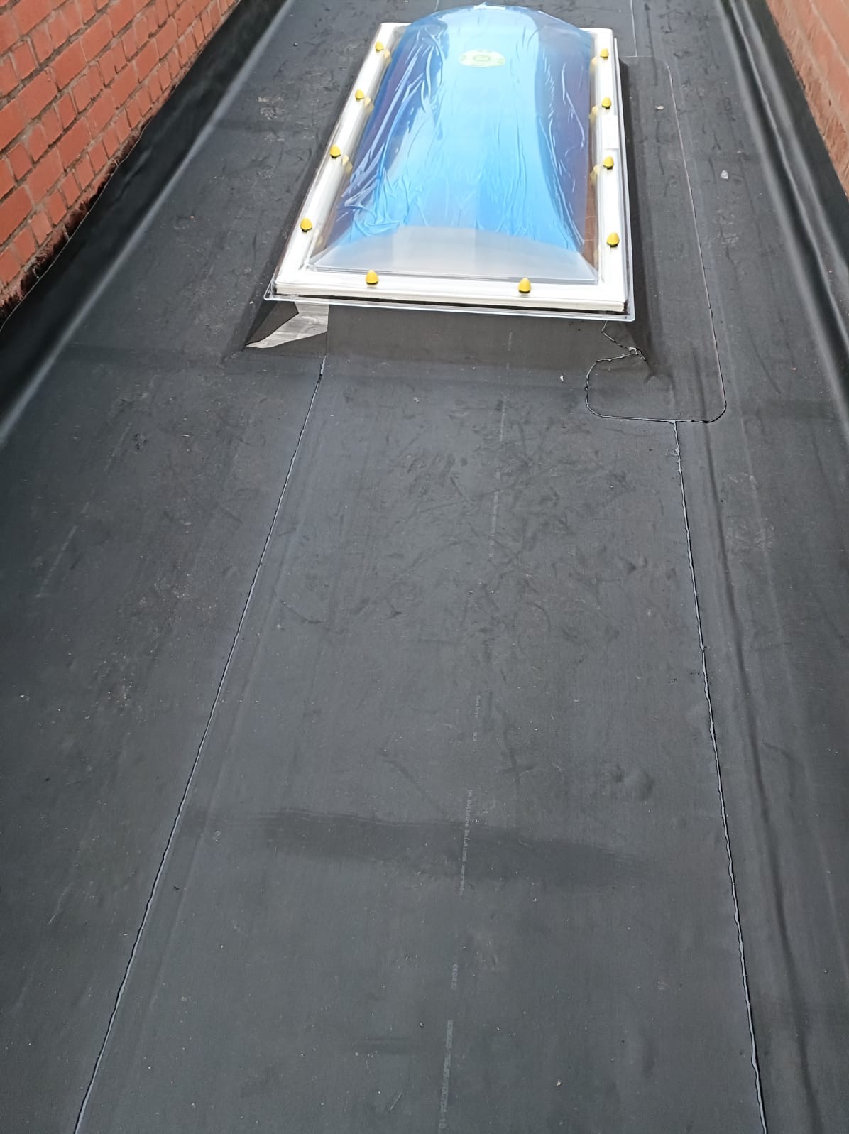 Dach pokryty czarną papą z wbudowanym świetlikiem dachowym zabezpieczonym folią, widok z góry. Detal łączenia papy przy świetliku.