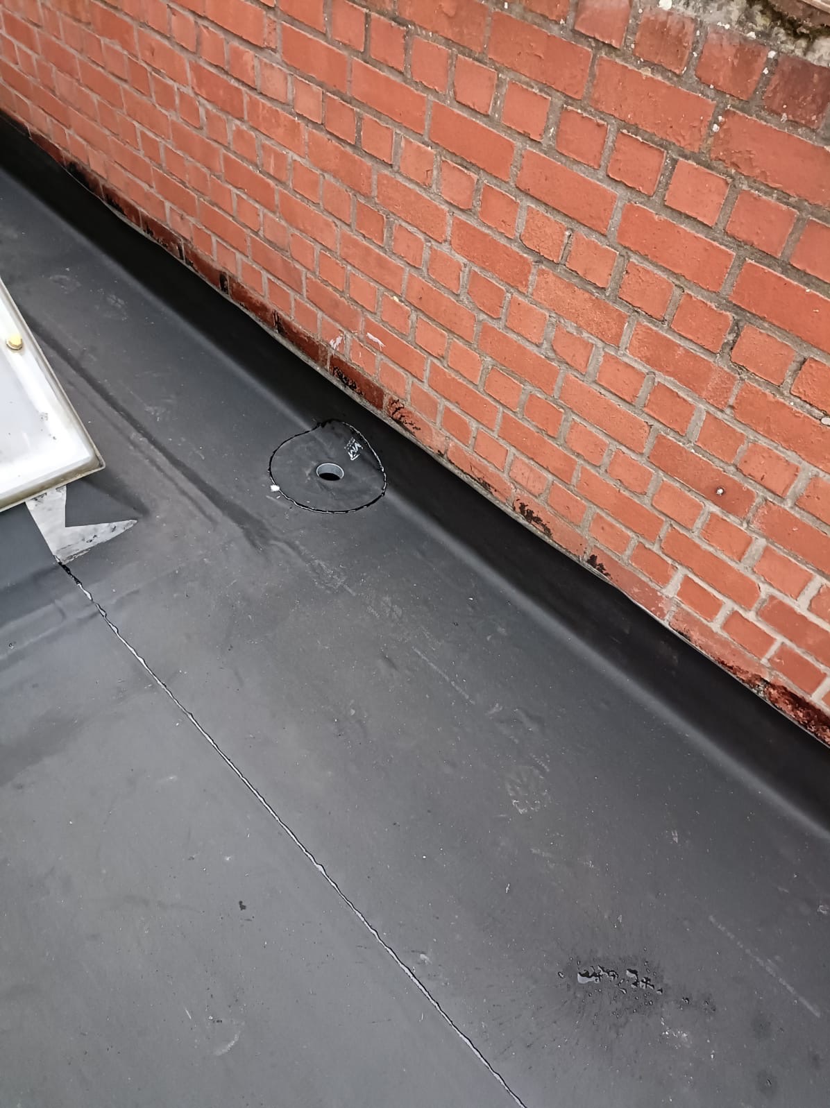 Płaski dach pokryty czarną membraną bitumiczną, przylegający do ceglanej ściany. Widoczny odpływ wody i pęknięcie na powierzchni dachu.