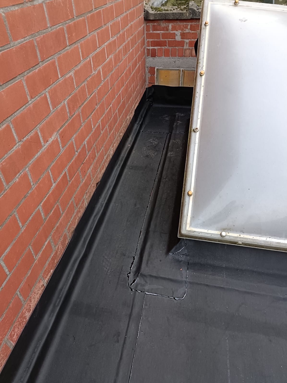 Fragment dachu pokrytego czarną papą z widocznymi pęknięciami i łatami, przylegającego do ceglanej ściany. W tle element świetlika dachowego.
