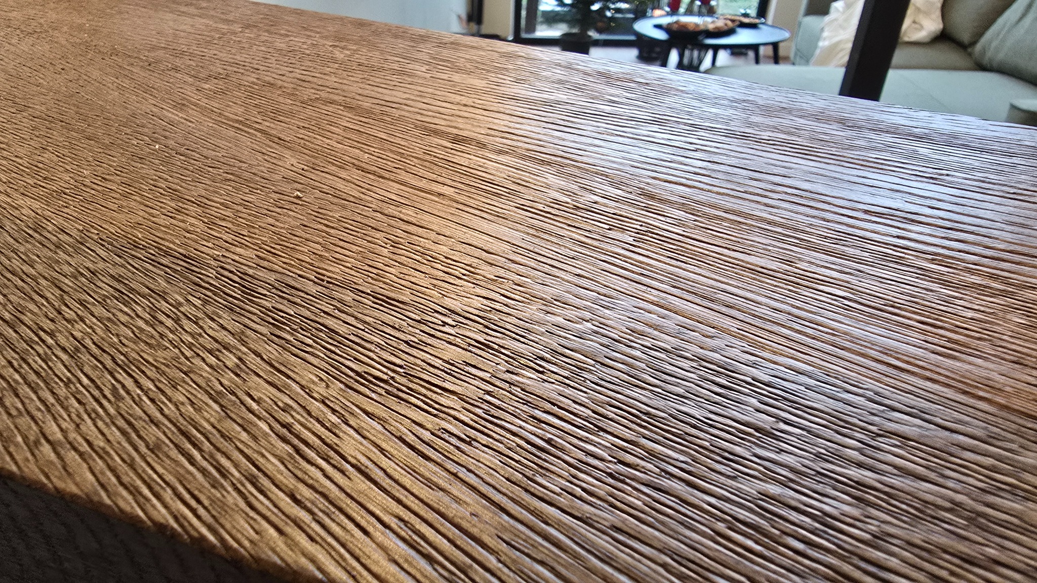 Teksturowany blat z wyraźnym usłojeniem, ciepłe odcienie brązu, widok z bliska, w tle fragment wnętrza z sofą i stolikiem.
