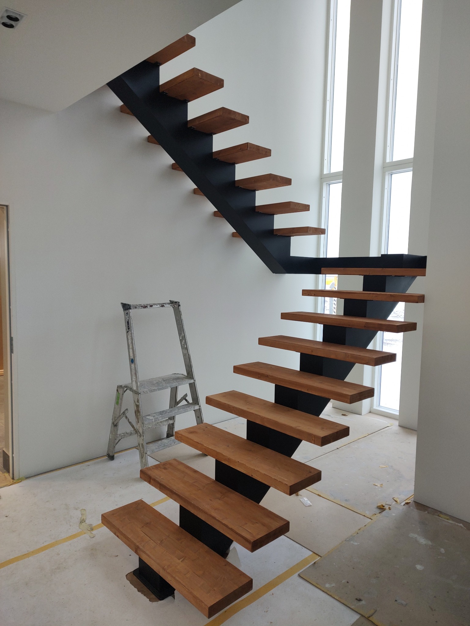 Nowoczesne schody wspornikowe z drewnianymi stopniami i czarną metalową konstrukcją, widoczne z bliska, obok drabina. Minimalistyczny design w jasnym wnętrzu.