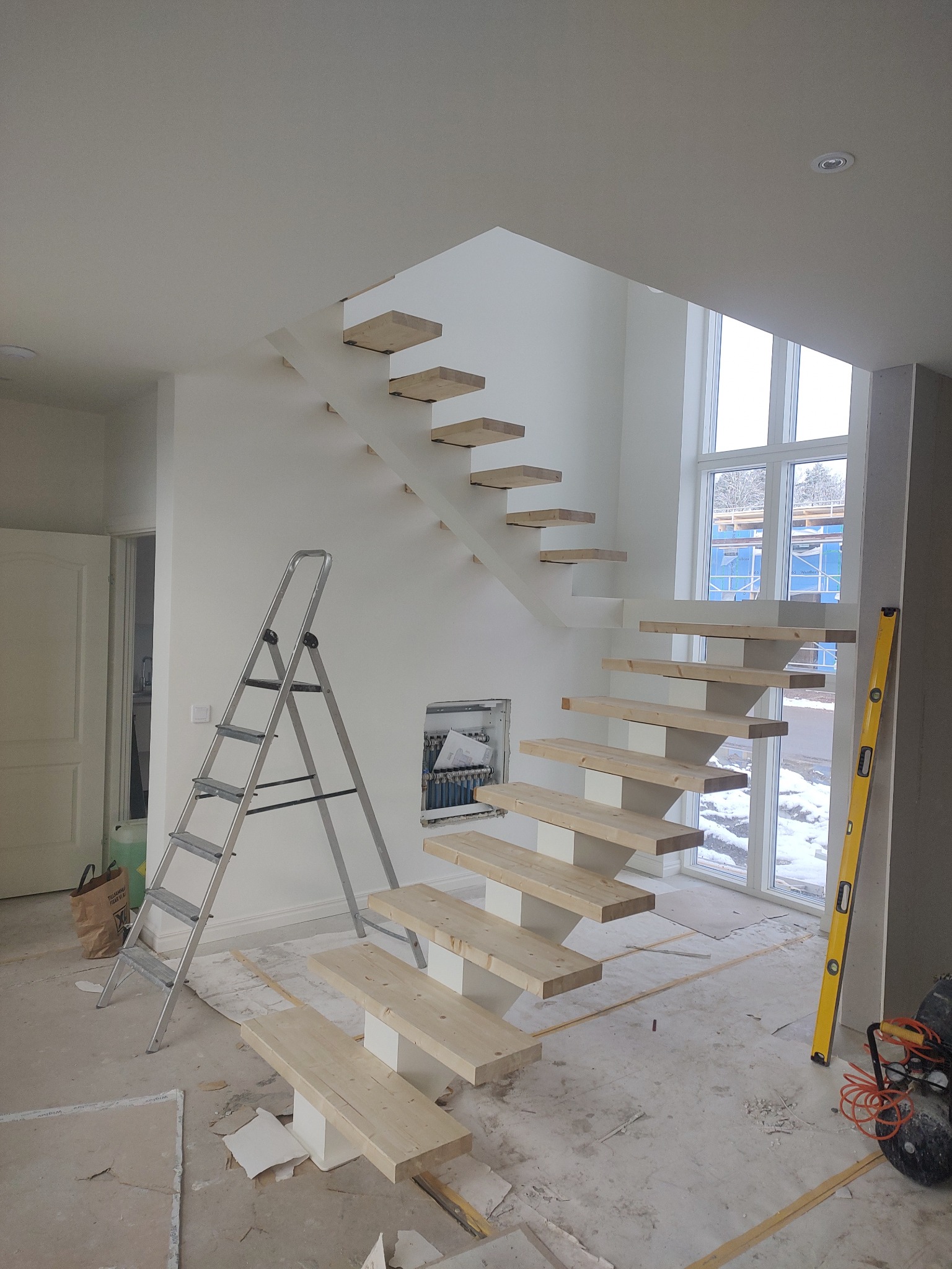 Montaż schodów dywanowych z drewnianymi stopniami. Widoczna drabina, poziomica i okno z widokiem na zimowy krajobraz. Surowy stan wykończenia wnętrza.