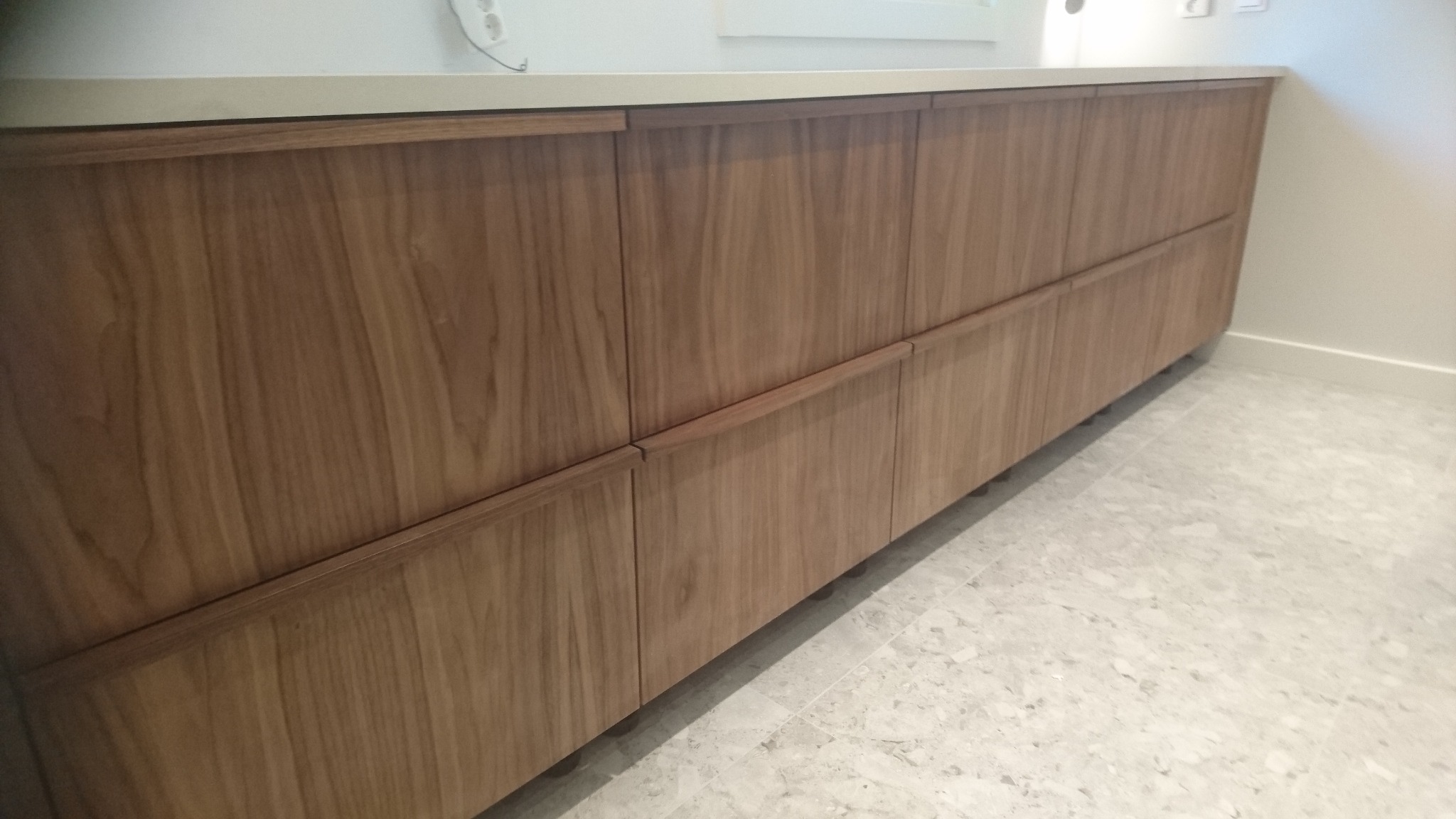 Front kuchni na wymiar z drewna orzechowego z subtelnymi, minimalistycznymi uchwytami, ustawionej na tle jasnej podłogi z terazzo.