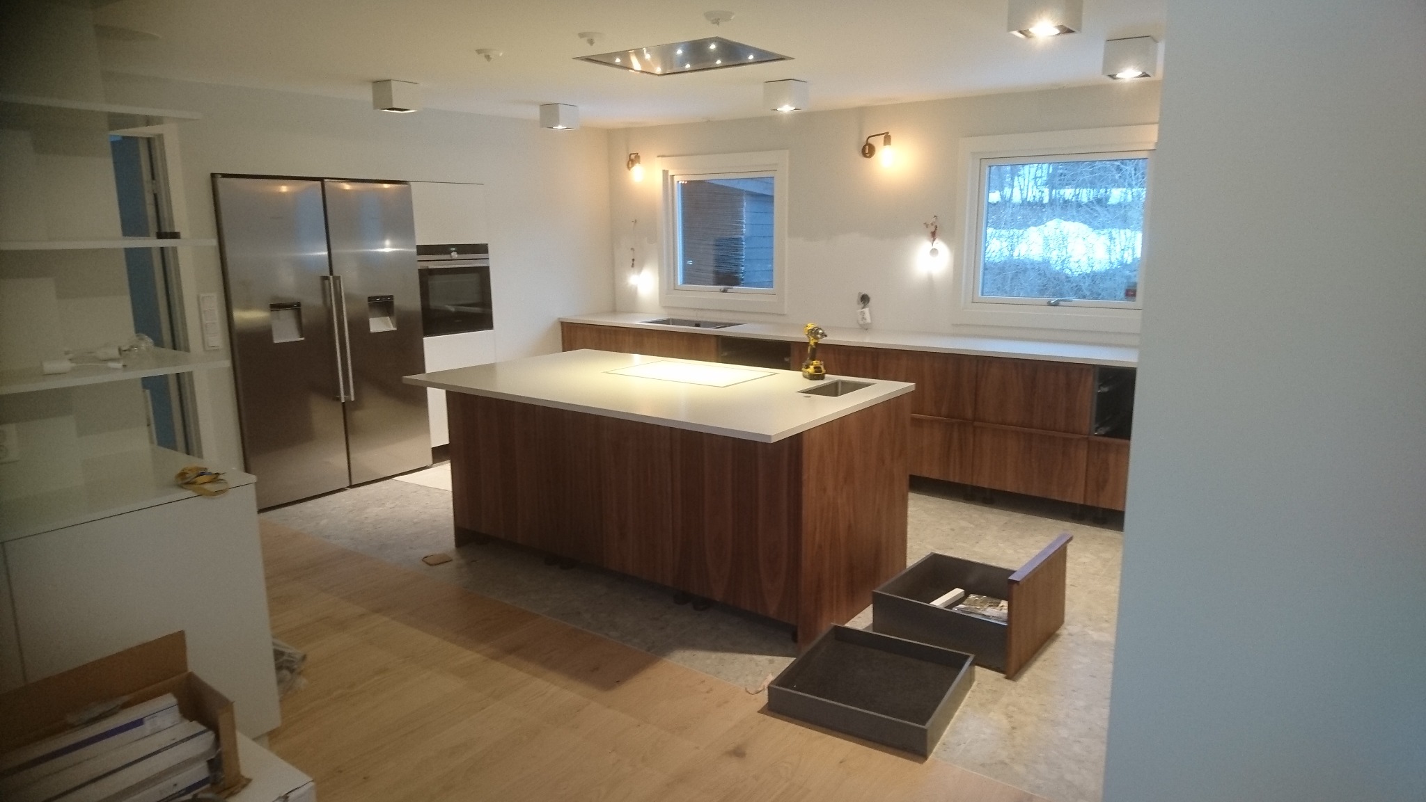 Nowoczesna kuchnia z wyspą i drewnianymi frontami szafek. Widoczne okna i sprzęt AGD. Na podłodze panele. Częściowo widoczne szuflady pod wyspą.