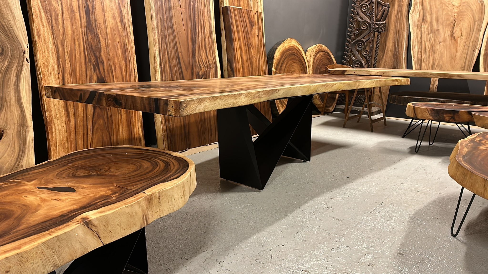Stół z blatem z egzotycznego drewna na czarnych, metalowych nogach. W tle deski i stoliki kawowe z tego samego materiału. Unikalny wzór słojów drewna.