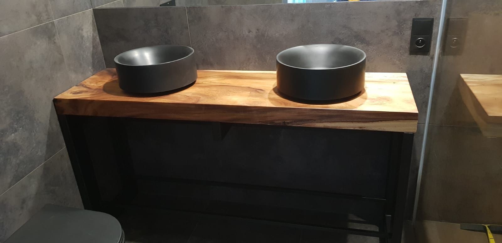 Nowoczesna łazienka z drewnianym blatem i dwiema czarnymi umywalkami typu vessel. Minimalistyczny design, szare płytki i subtelne oświetlenie.