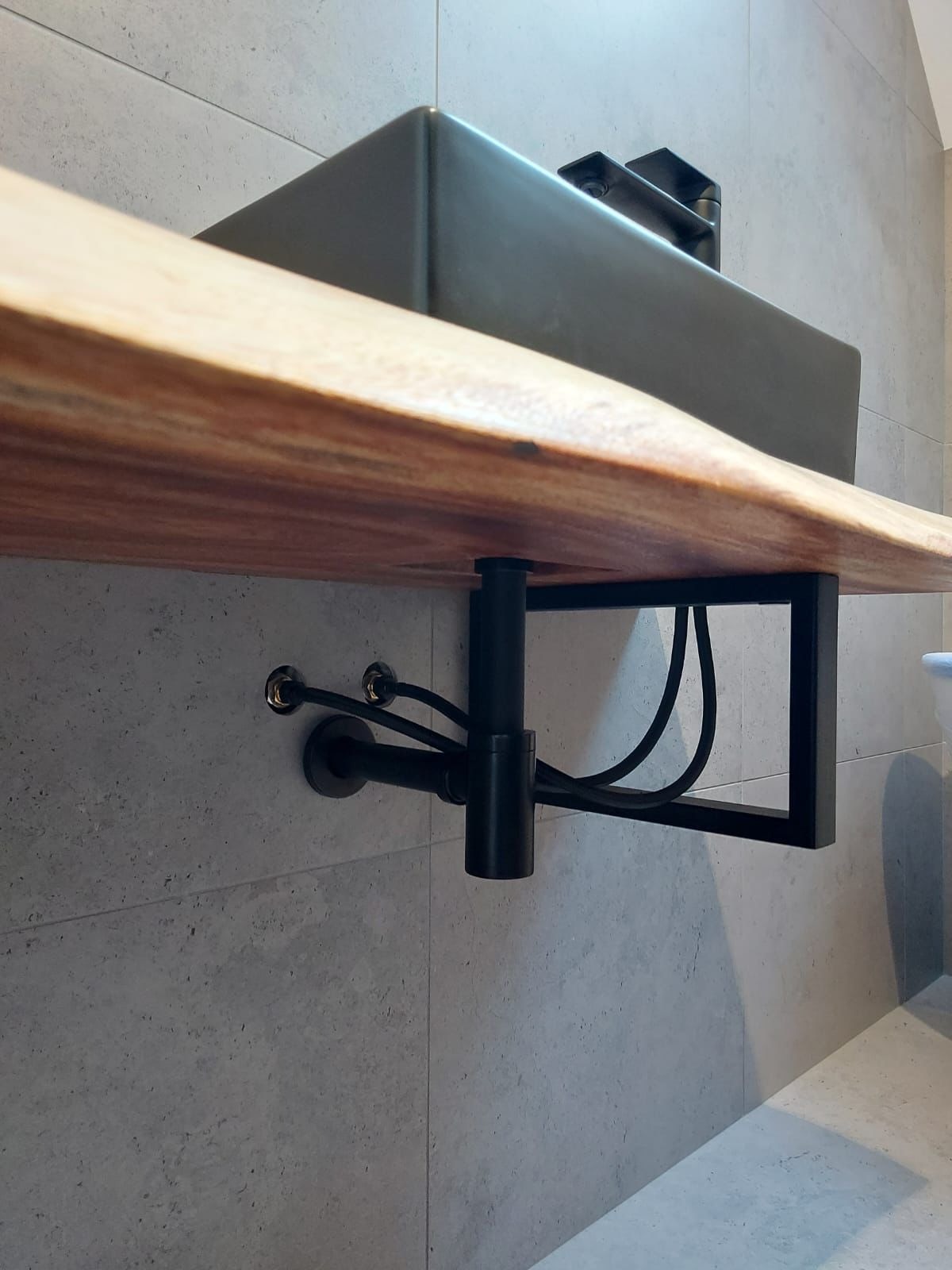 Drewniany blat łazienkowy z umywalką o surowym wykończeniu, wsparty na czarnym, metalowym stelażu. Widoczne przyłącza wodne i kanalizacyjne na tle szarych płytek.