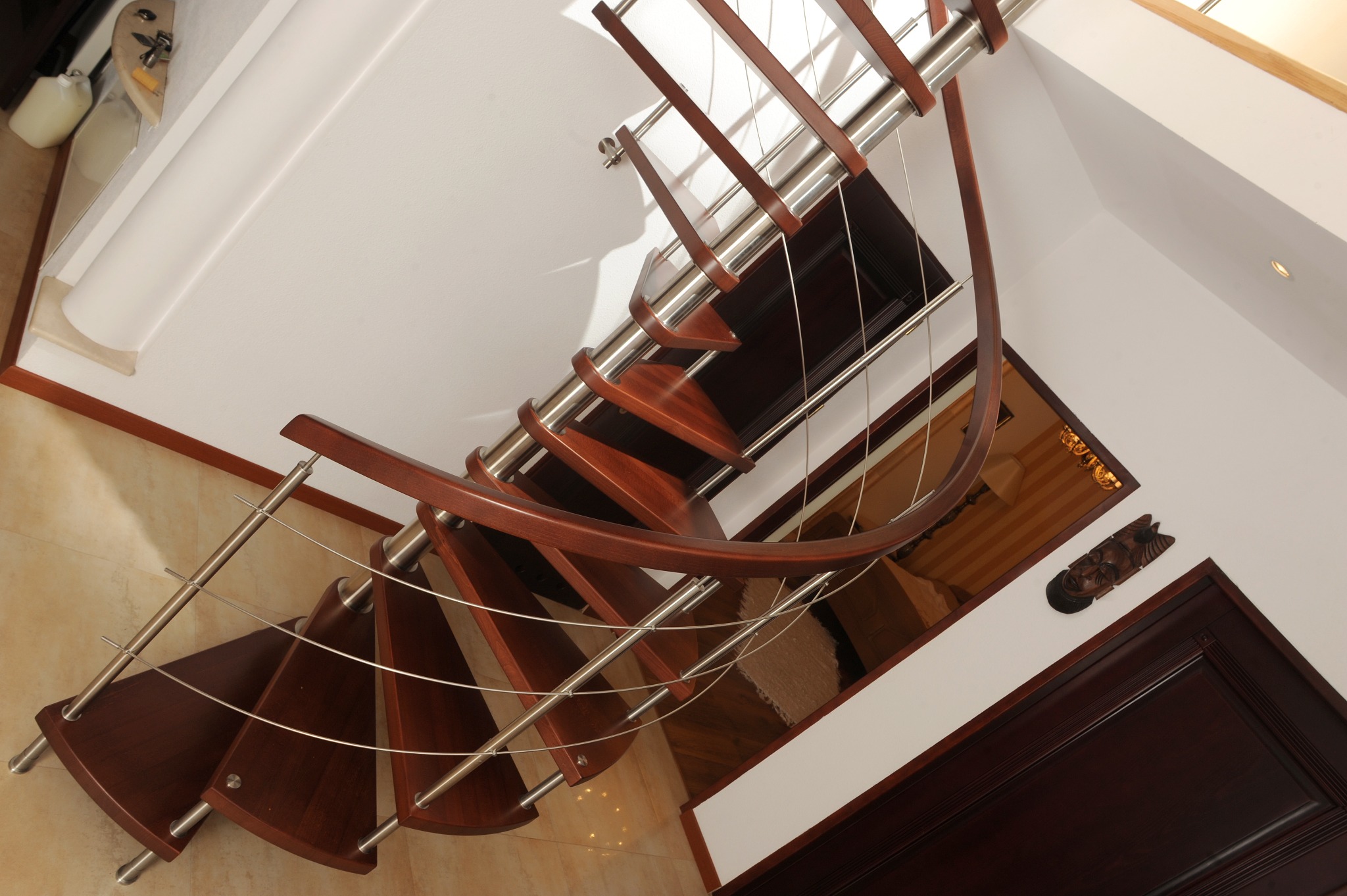 Spiralne schody z ciemnego drewna i metalu, widziane z dołu, z balustradami z cienkich prętów. W tle widoczne drzwi i dekoracja na ścianie.