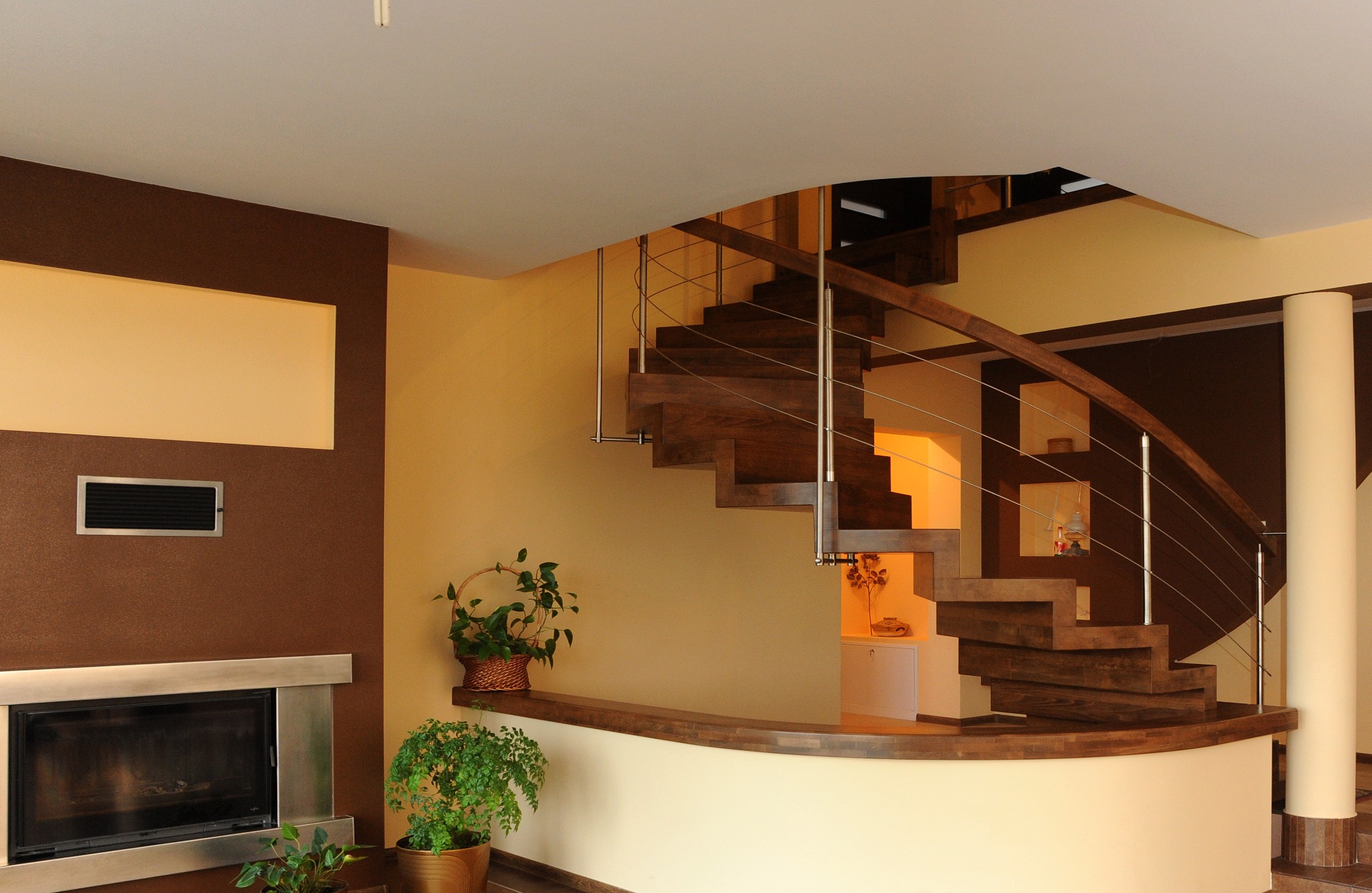 Eleganckie, drewniane schody spiralne z metalowymi balustradami w jasnym wnętrzu. Widoczne rośliny doniczkowe i kominek z metalowym obramowaniem.